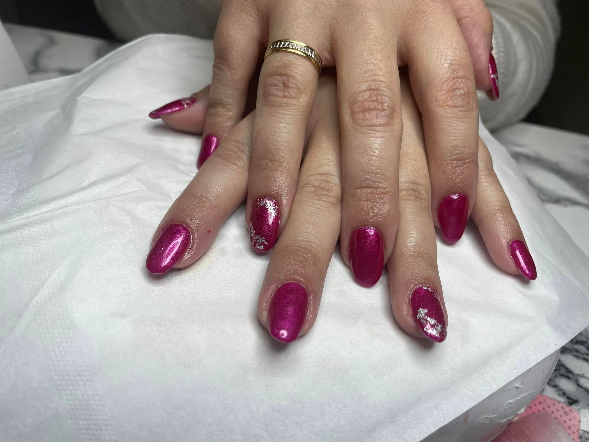 Manos con uñas ovaladas de color magenta, algunas con detalles plateados. Un anillo de oro en un dedo.