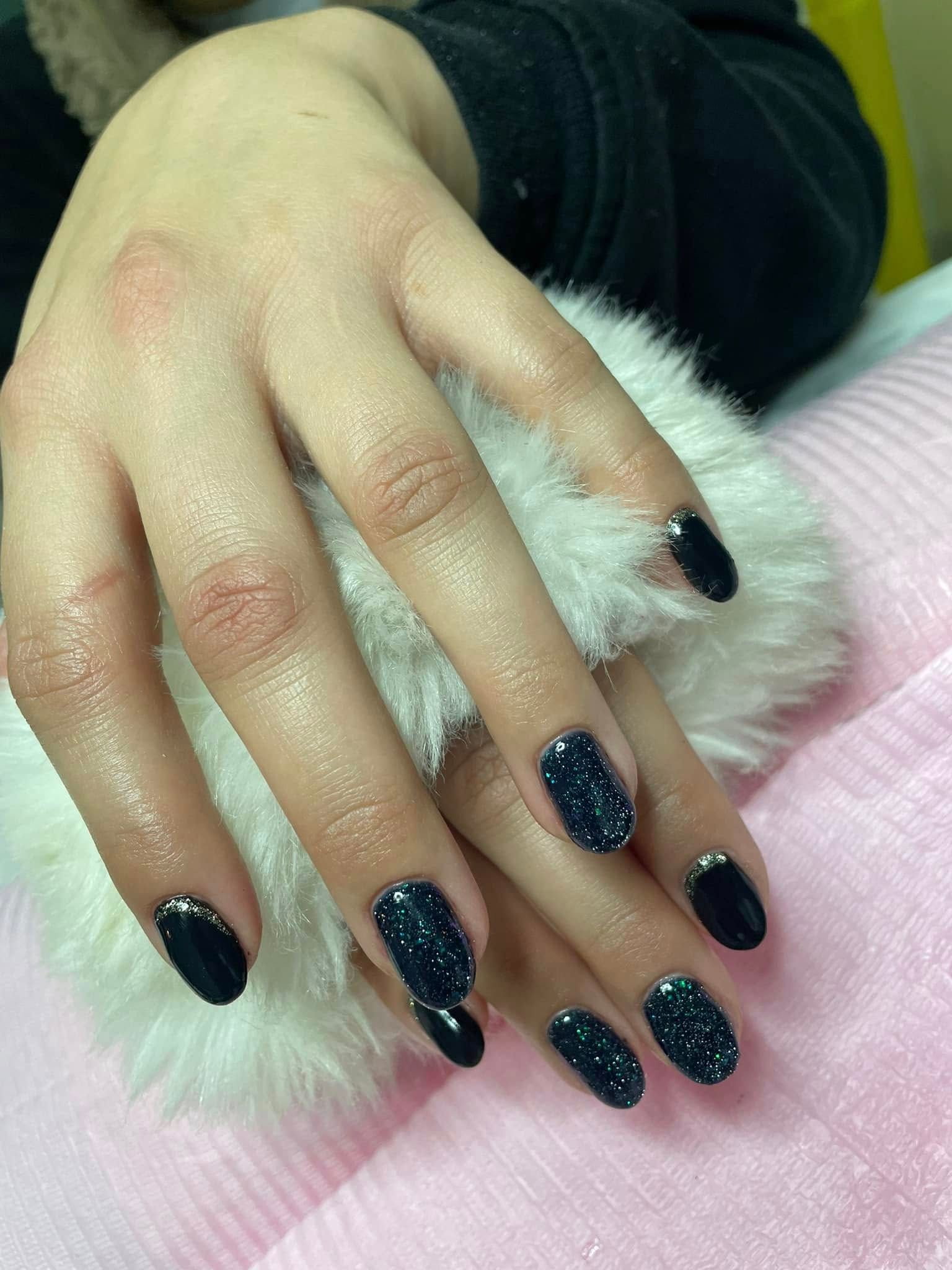 Manos con esmalte de uñas azul oscuro y detalles de brillo, descansando sobre una superficie blanca y esponjosa, fondo rosa.