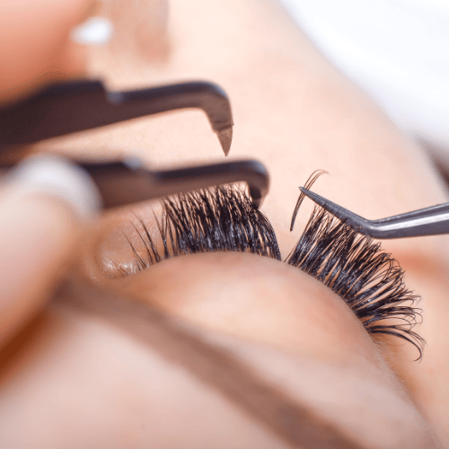 Aplicación de extensiones de pestañas; pinzas en uso. Primer plano de un ojo con pestañas oscuras y piel clara.