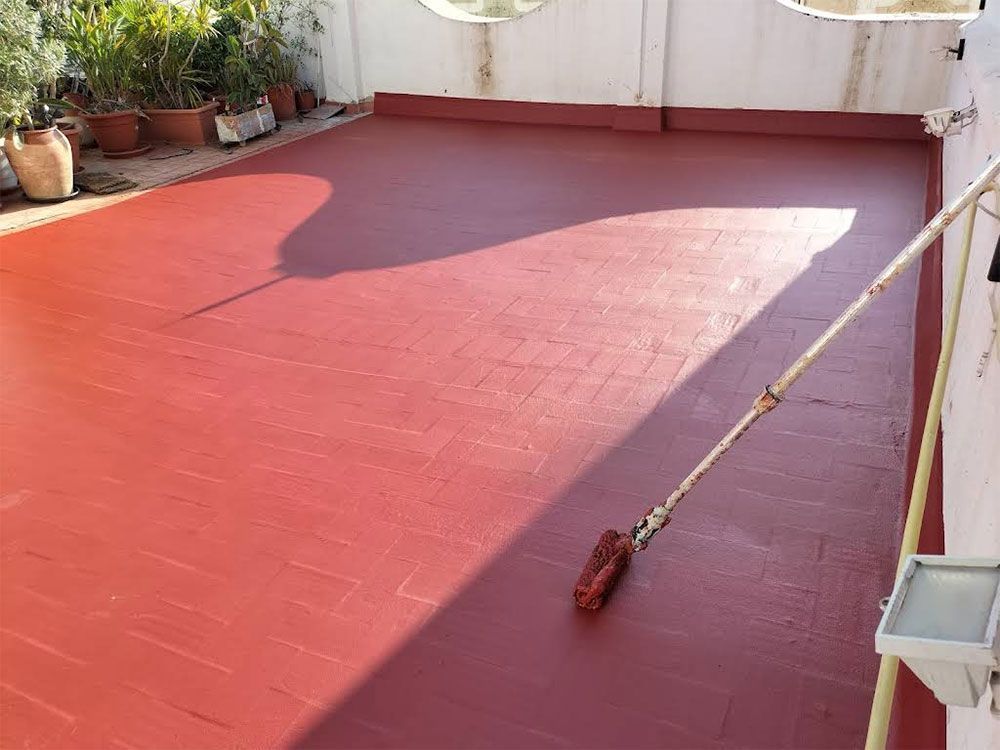 Una terraza en la azotea pintada de color marrón rojizo con un rodillo de pintura en el suelo, al lado de una pared blanca y plantas en macetas.