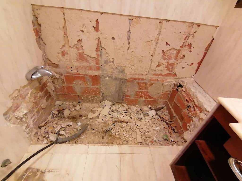 Una pared y un piso de baño con ladrillos a la vista y escombros de un área de ducha o bañera parcialmente demolida.