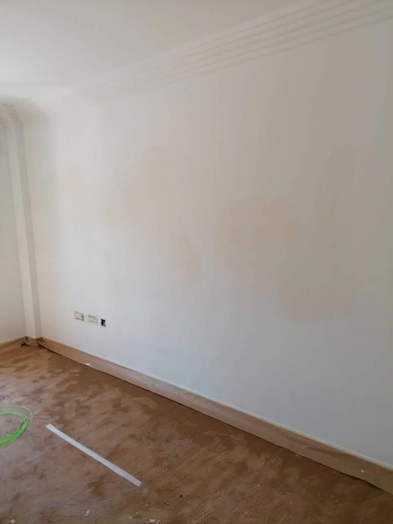 Una pared blanca sencilla en una habitación con alfombra de color claro, que muestra una ligera decoloración de la pintura y enchufes eléctricos.