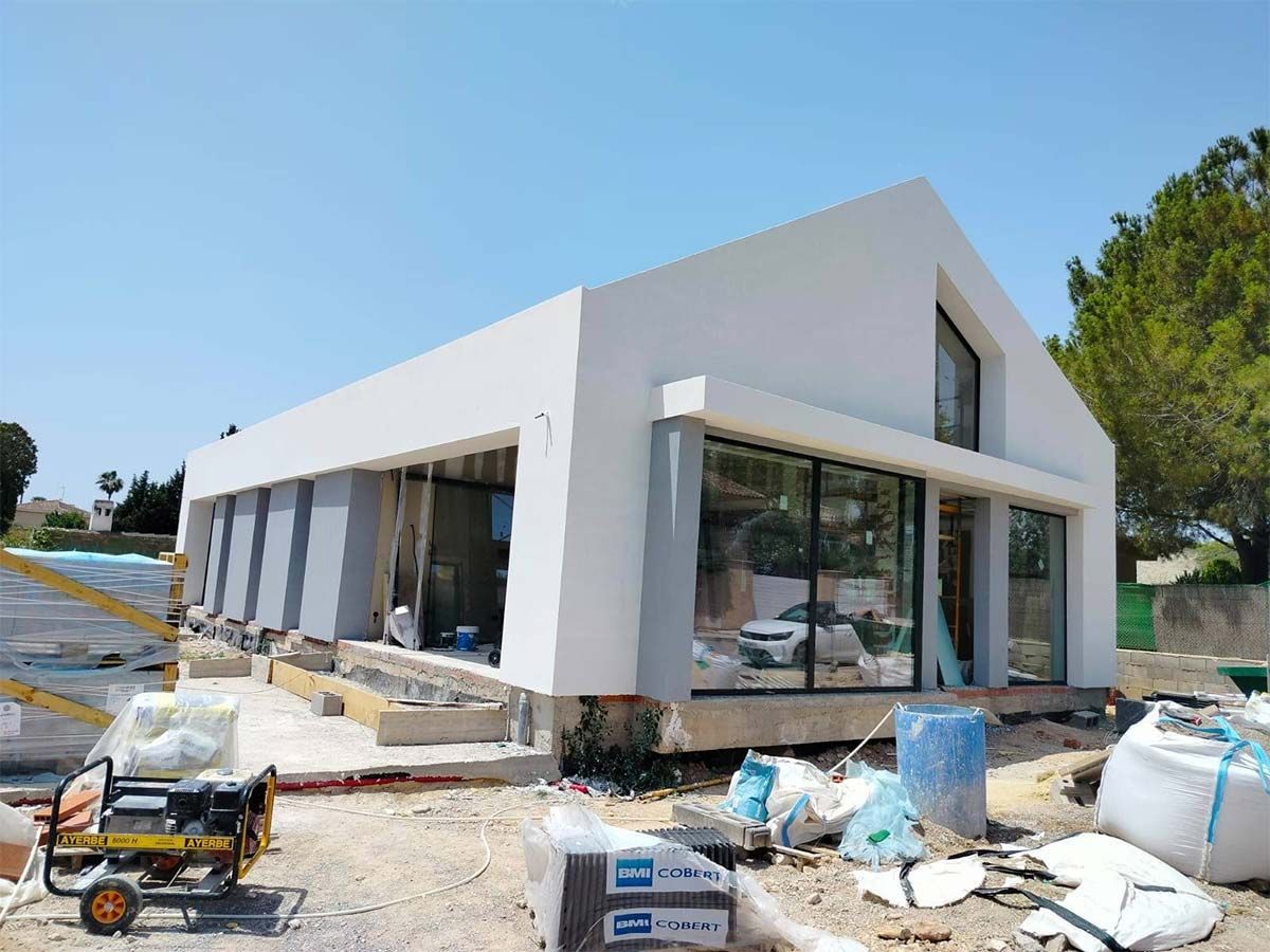 Un edificio blanco, moderno, con techo a dos aguas, en construcción, con grandes ventanales y equipos de construcción en el lugar.