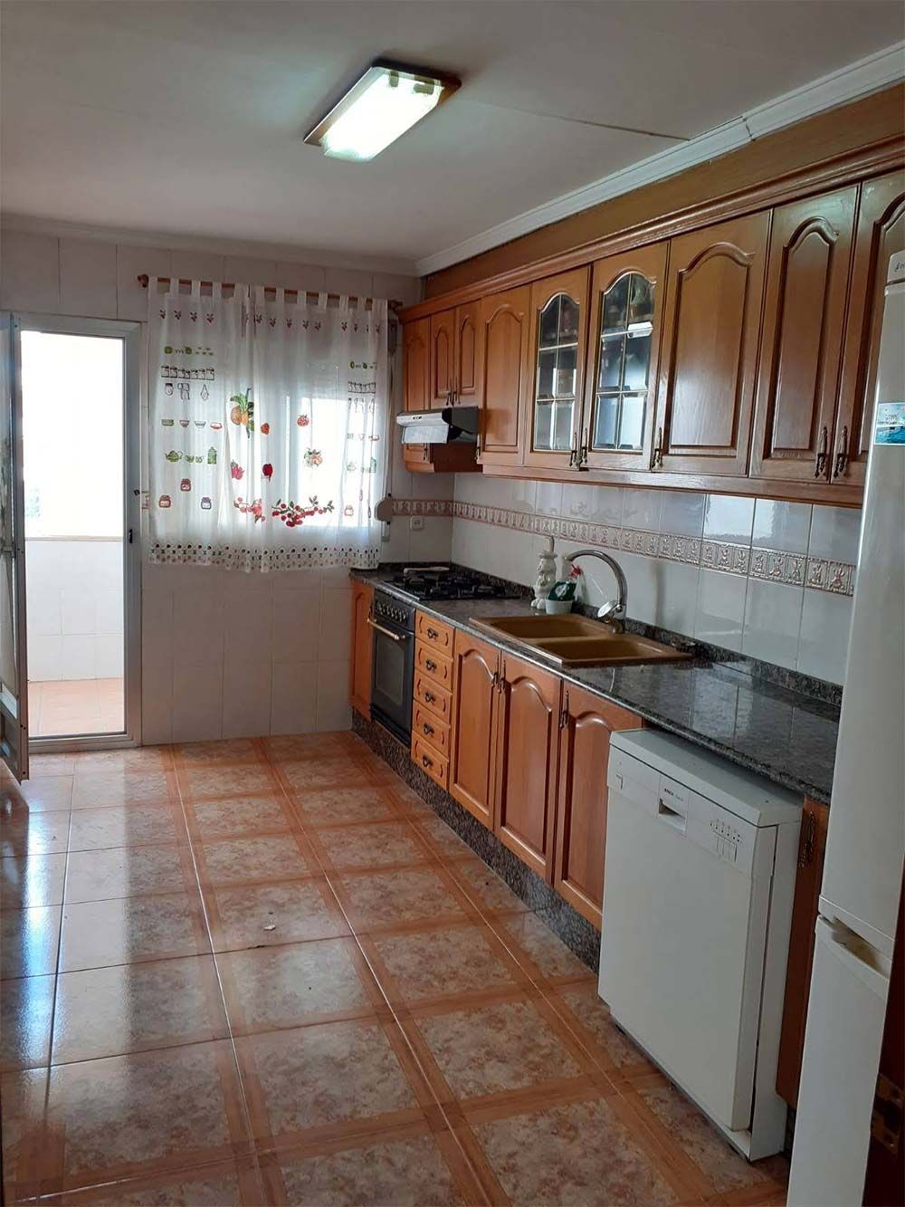 Una cocina estrecha con gabinetes de madera clara, encimeras oscuras, pisos de baldosas y una ventana con cortinas estampadas.