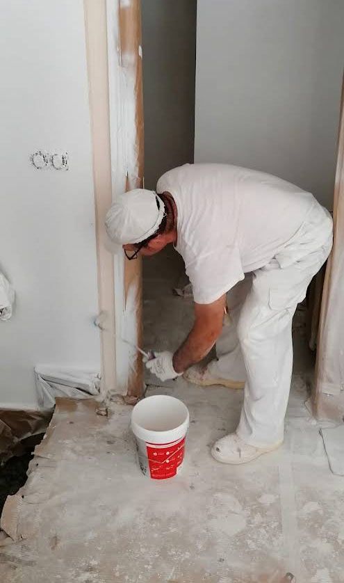 Un trabajador vestido completamente de blanco pinta los bordes de una puerta en una habitación en construcción con un balde de pintura blanca.