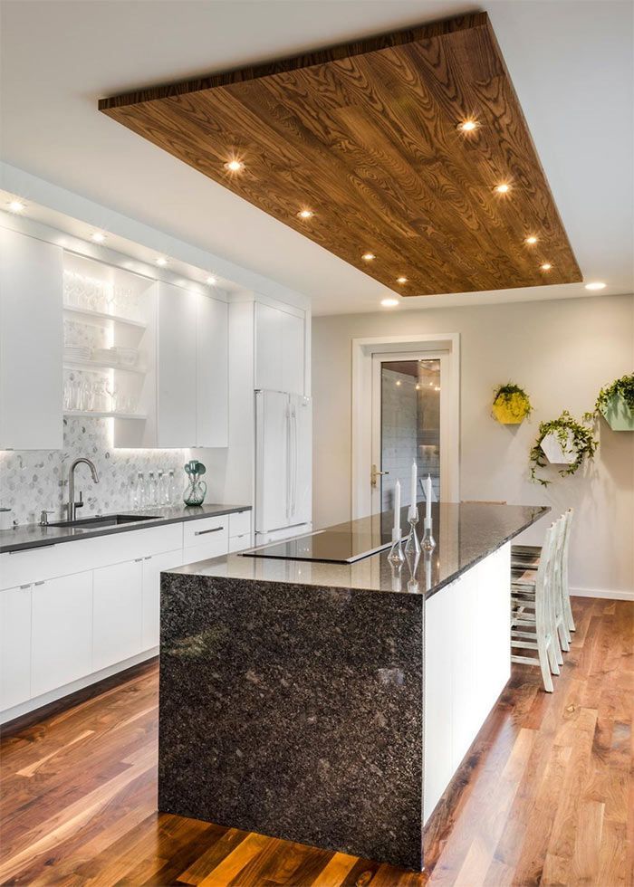 Una cocina moderna con gabinetes blancos, isla de granito oscuro, techo de tablones de madera y pisos de madera.