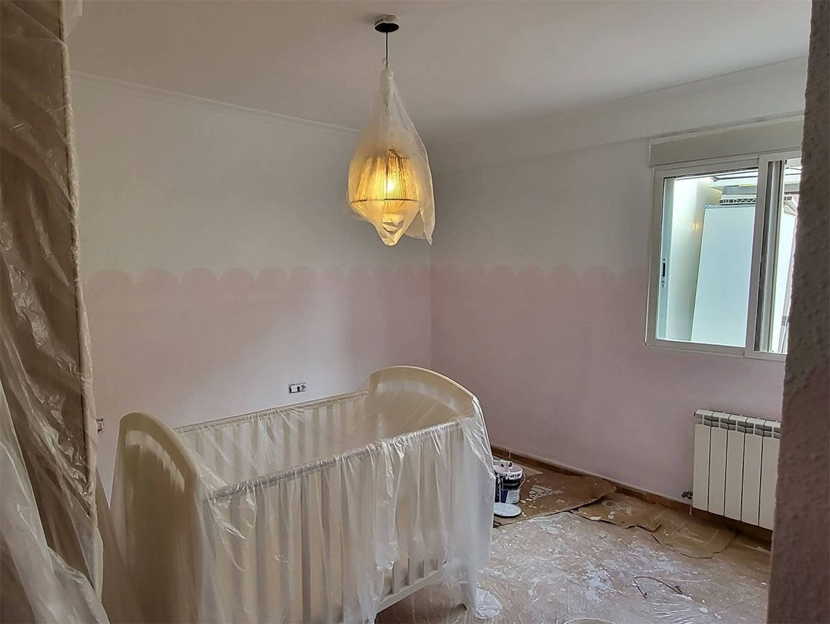 Una cuna blanca se encuentra en una habitación con paredes parcialmente pintadas de rosa, una lámpara colgante y escombros en el suelo.