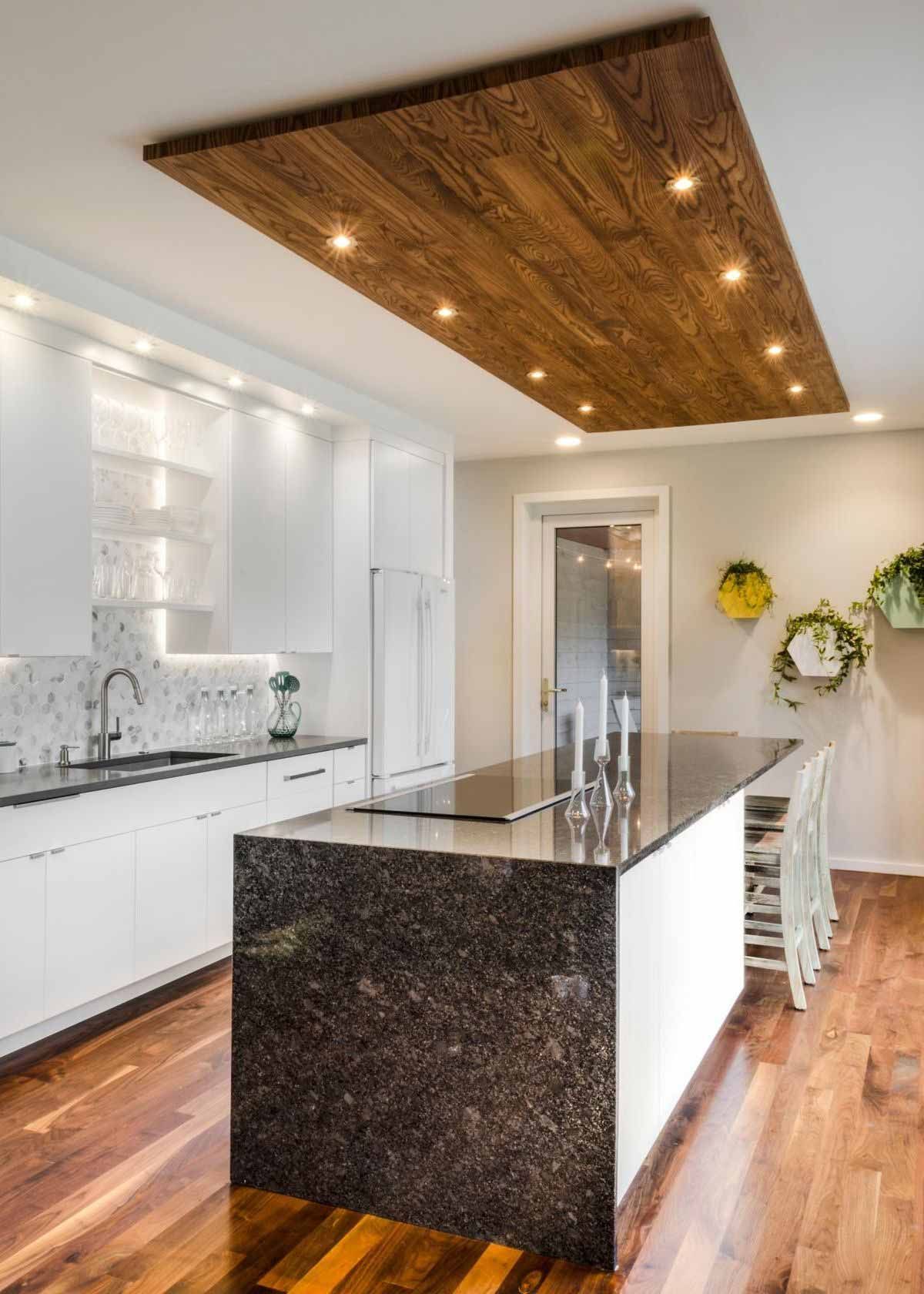 Una cocina con gabinetes blancos, una gran isla de granito oscuro y un distintivo techo de tablones de madera con focos.