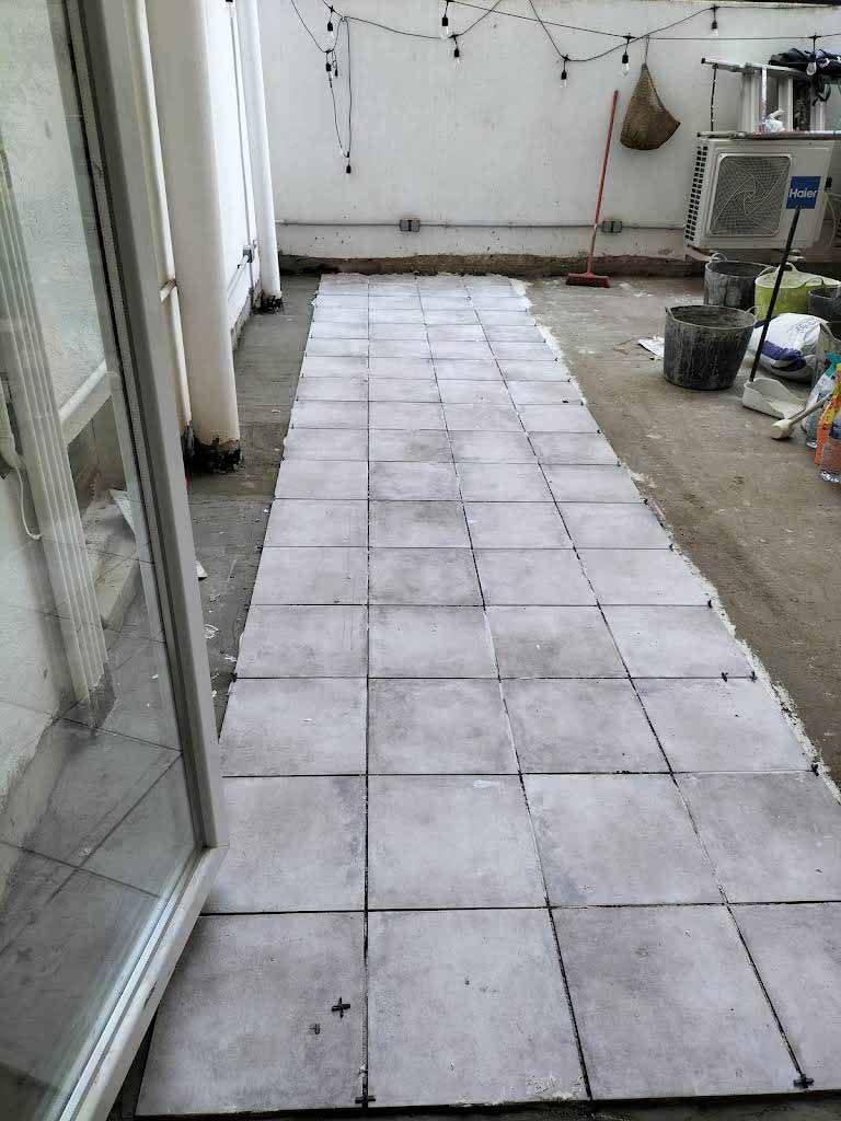 Se está colocando un camino de baldosas grises cuadradas en el piso de un patio al aire libre, con líneas de lechada oscuras visibles entre ellas.