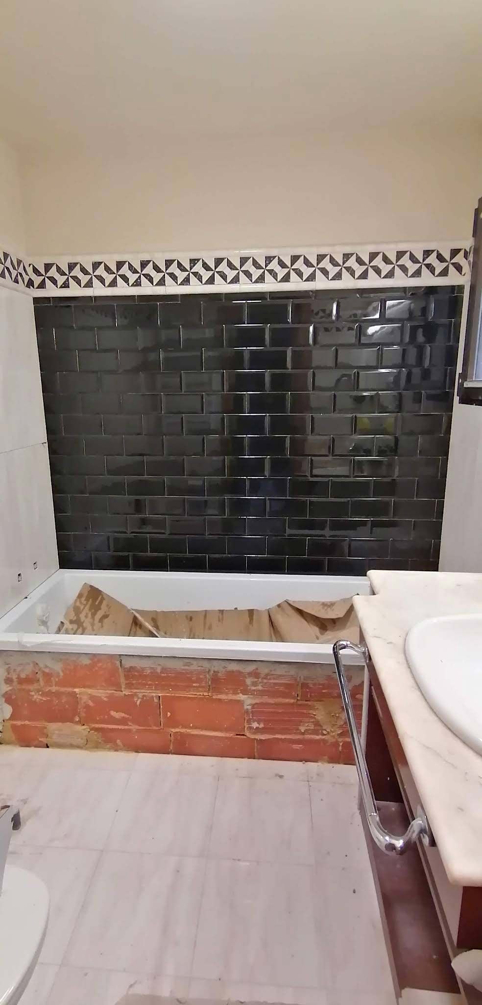 Una renovación de baño que incluye un revestimiento de bañera con azulejos negros tipo metro sobre una base de bañera de ladrillo sin terminar.