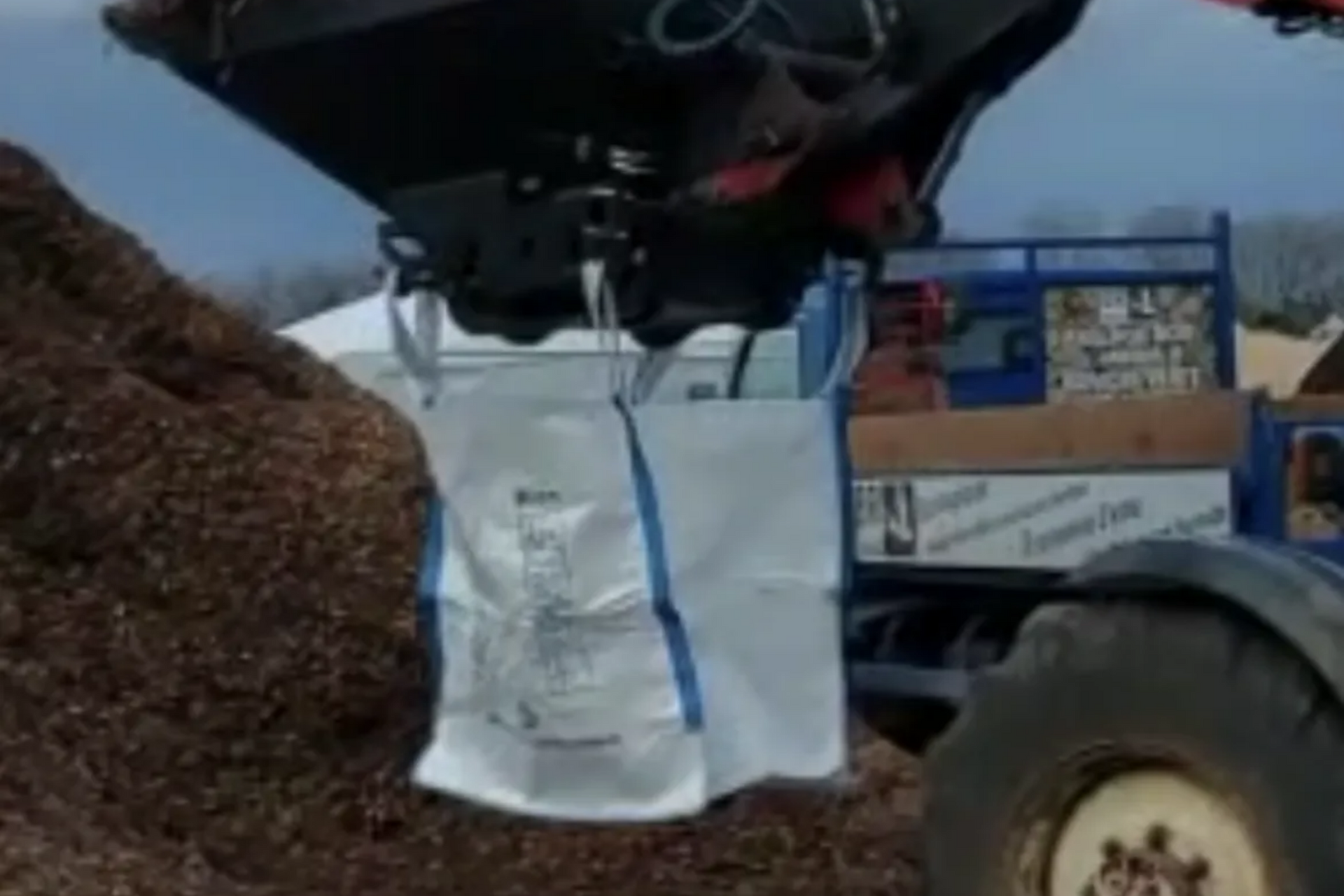 Une chargeuse soulève un grand sac blanc rempli de copeaux de bois.