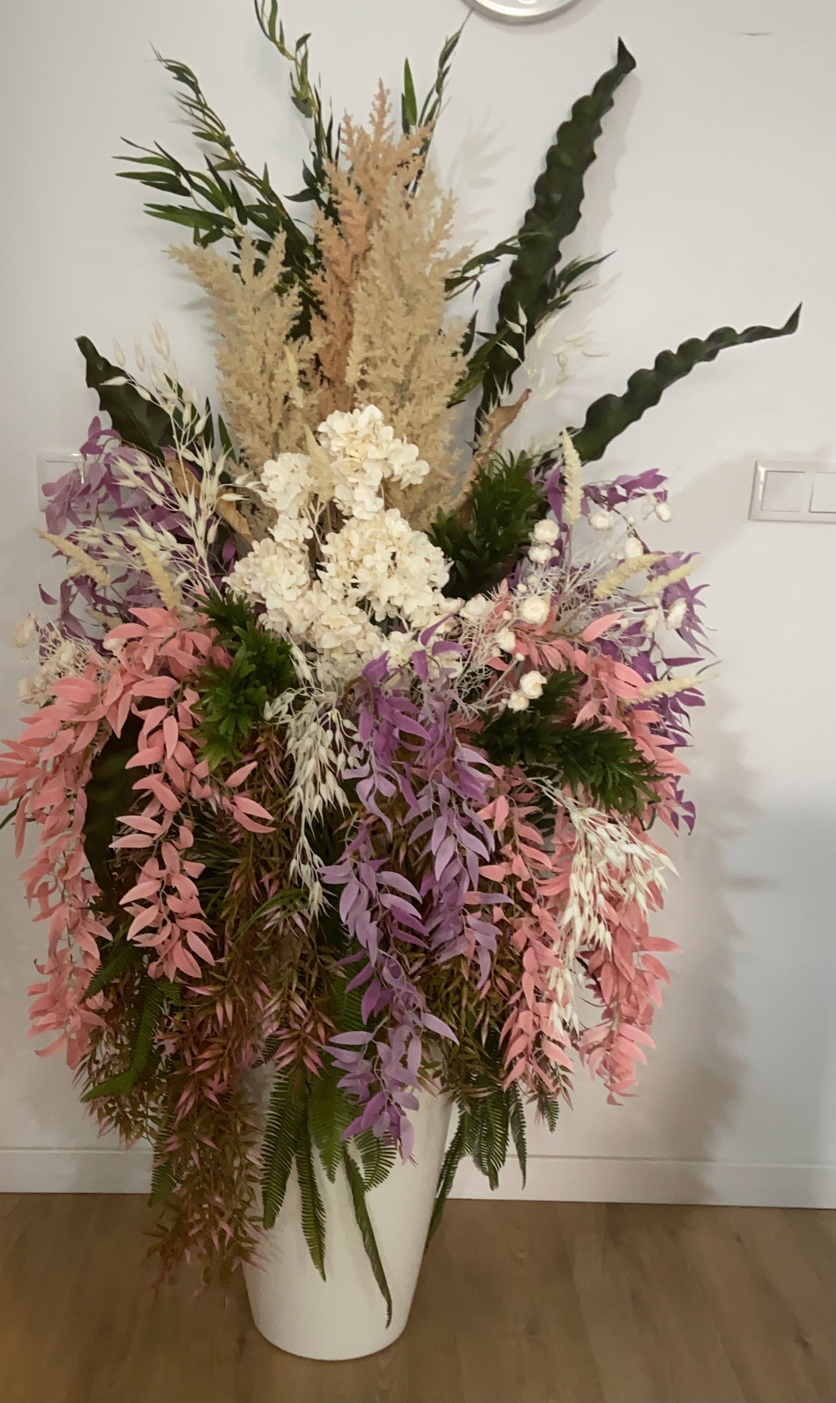 Arreglo floral grande y colorido en una maceta blanca contra una pared blanca y un piso de madera.