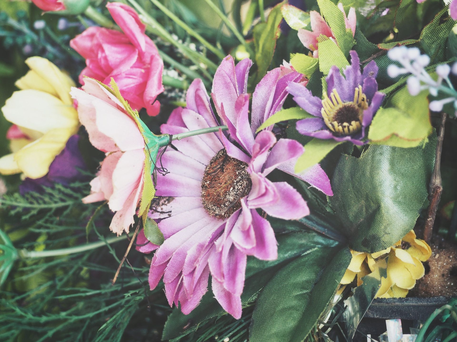 Primer plano de flores artificiales vibrantes, incluidas margaritas rosas y moradas, flores amarillas y hojas verdes.