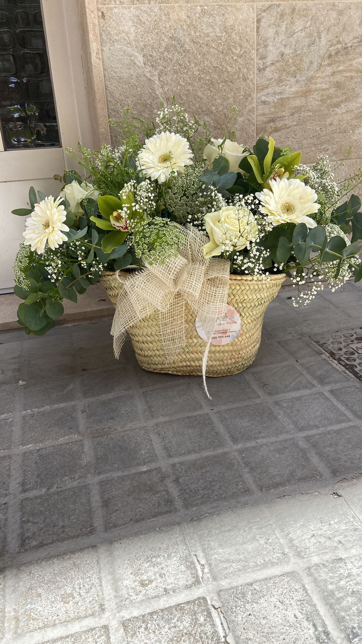 Cesta de flores blancas y vegetación con un lazo sobre un suelo de baldosas cerca de una puerta.