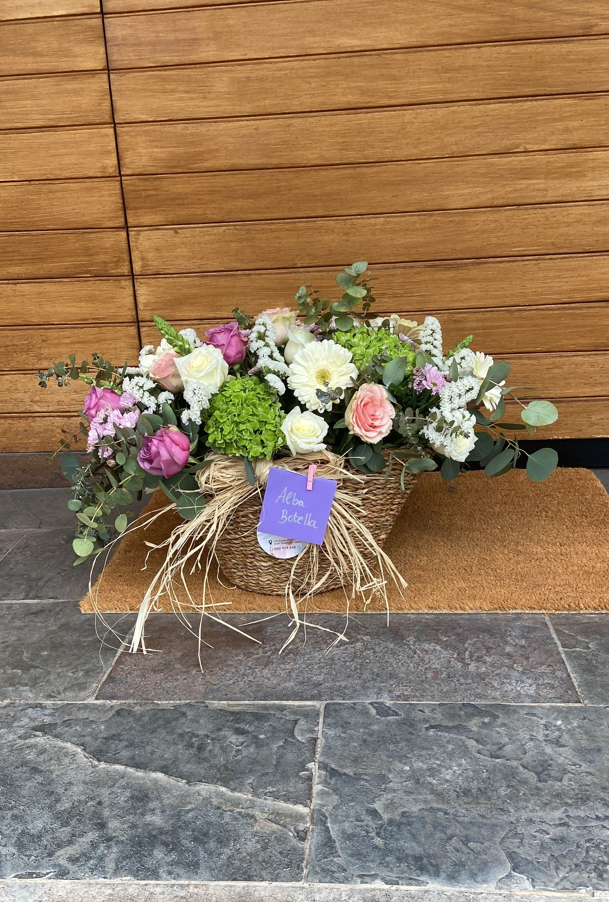 Cesta de flores blancas y vegetación con un lazo sobre un suelo de baldosas cerca de una puerta.