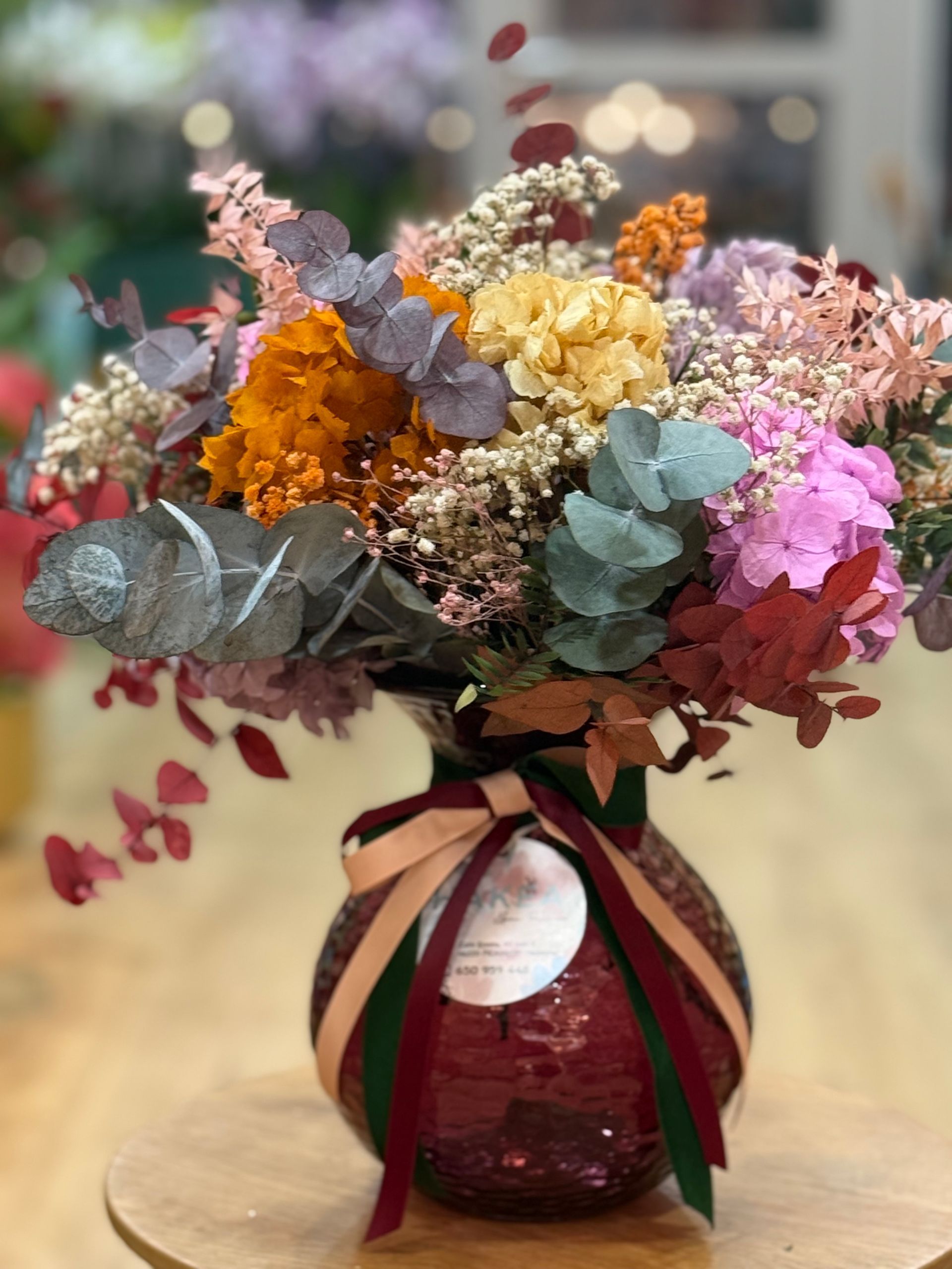 Ramo de flores secas de colores en un jarrón con lentejuelas rojas y cintas, sobre una superficie de madera.