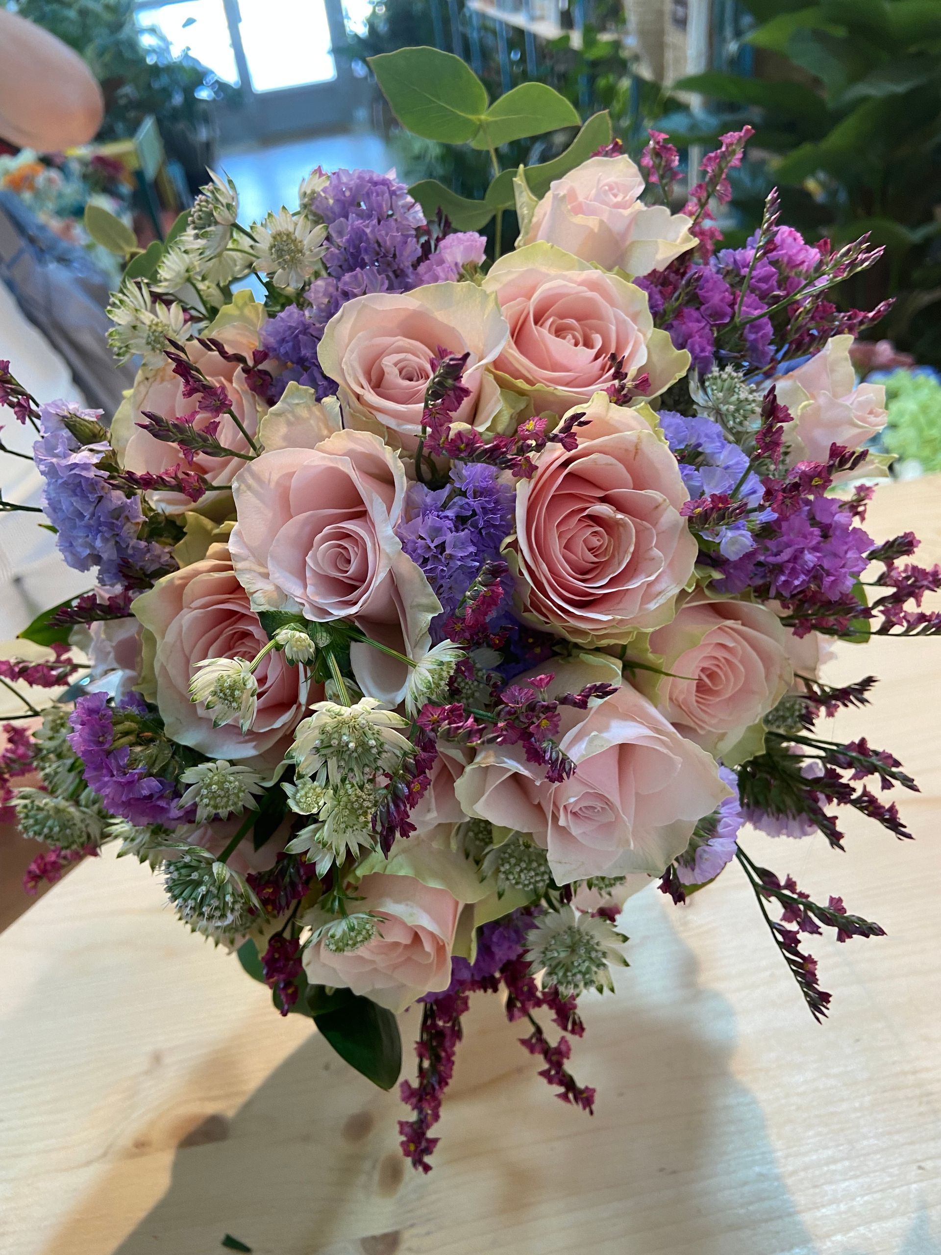 Ramo de rosas de color rosa claro, estátice morado y relleno verde sobre una superficie de madera.