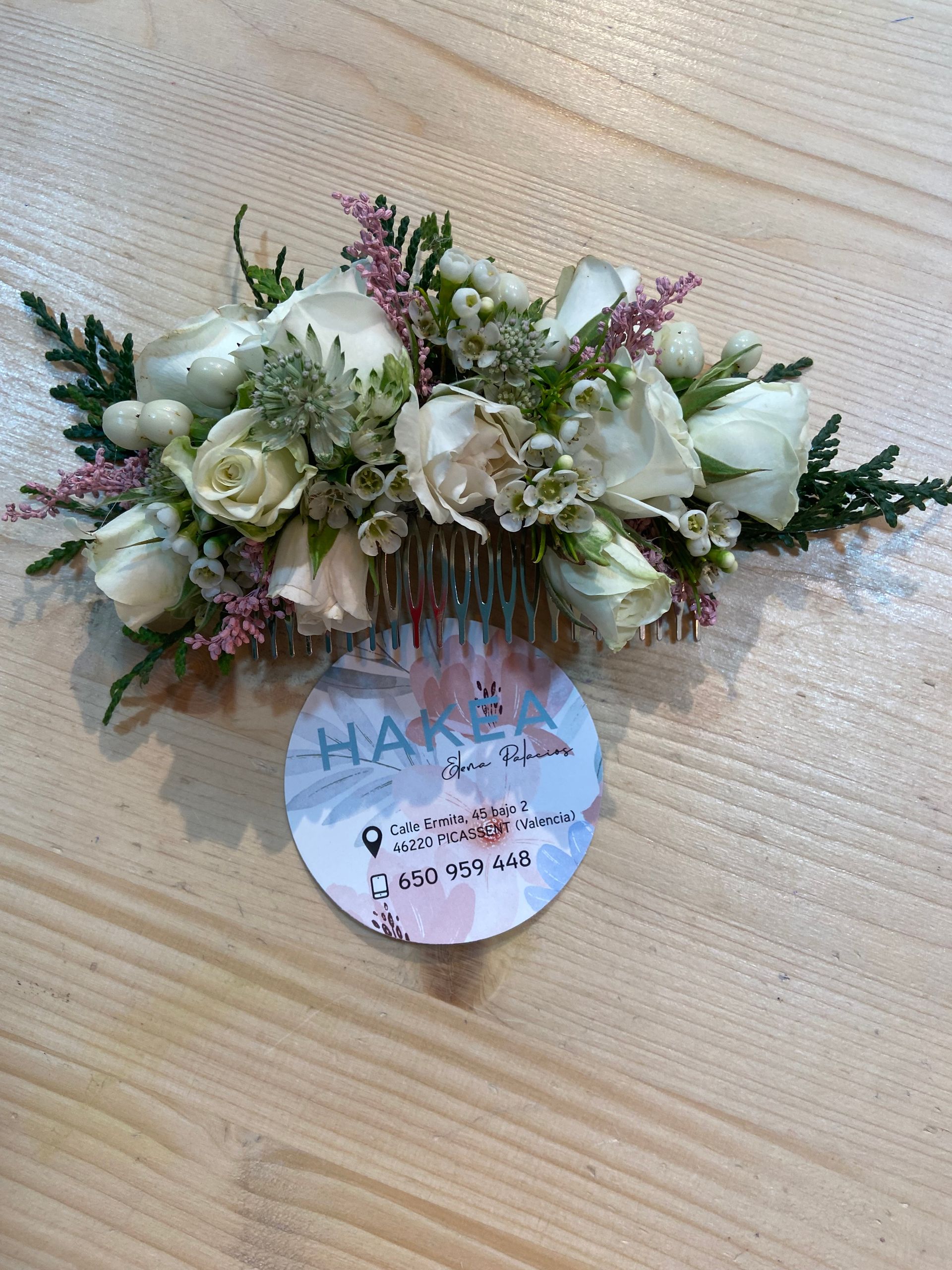 Peineta floral con rosas blancas y pequeñas flores rosas sobre una superficie de madera, con una tarjeta de presentación debajo.