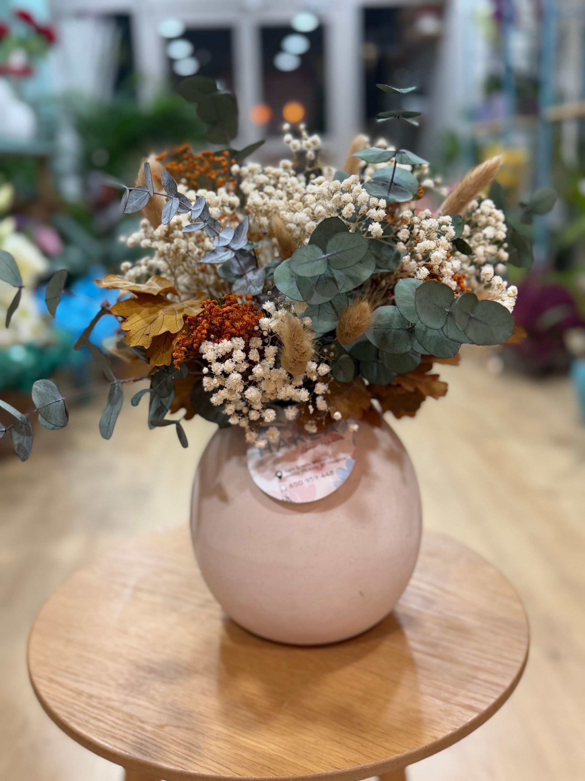 Arreglo floral en un jarrón esférico rosa claro sobre una mesa de madera. El ramo contiene flores secas y hojas de eucalipto.