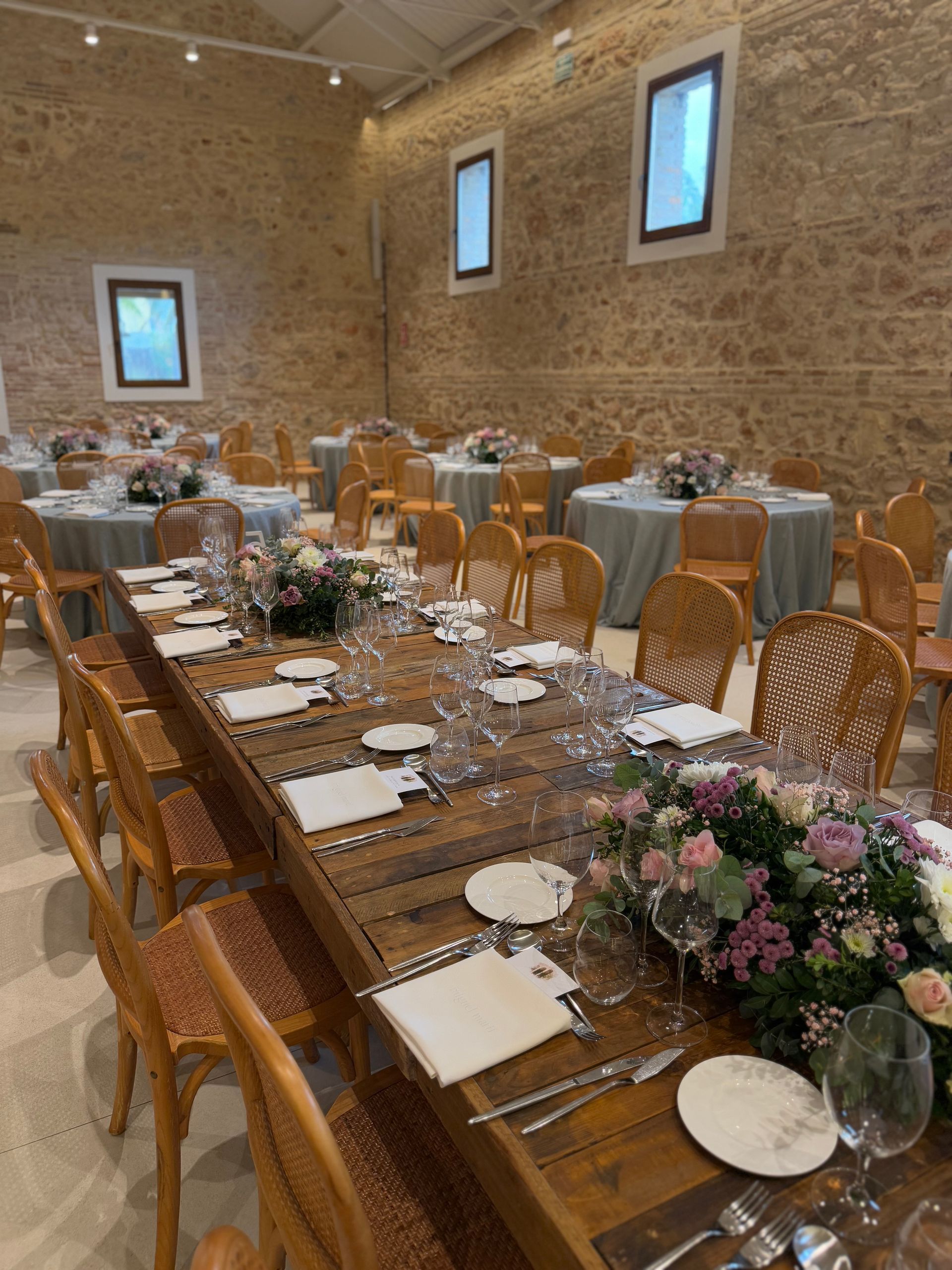 Salón de bodas con largas mesas de madera preparadas para la cena y mesas redondas al fondo. Paredes de piedra.