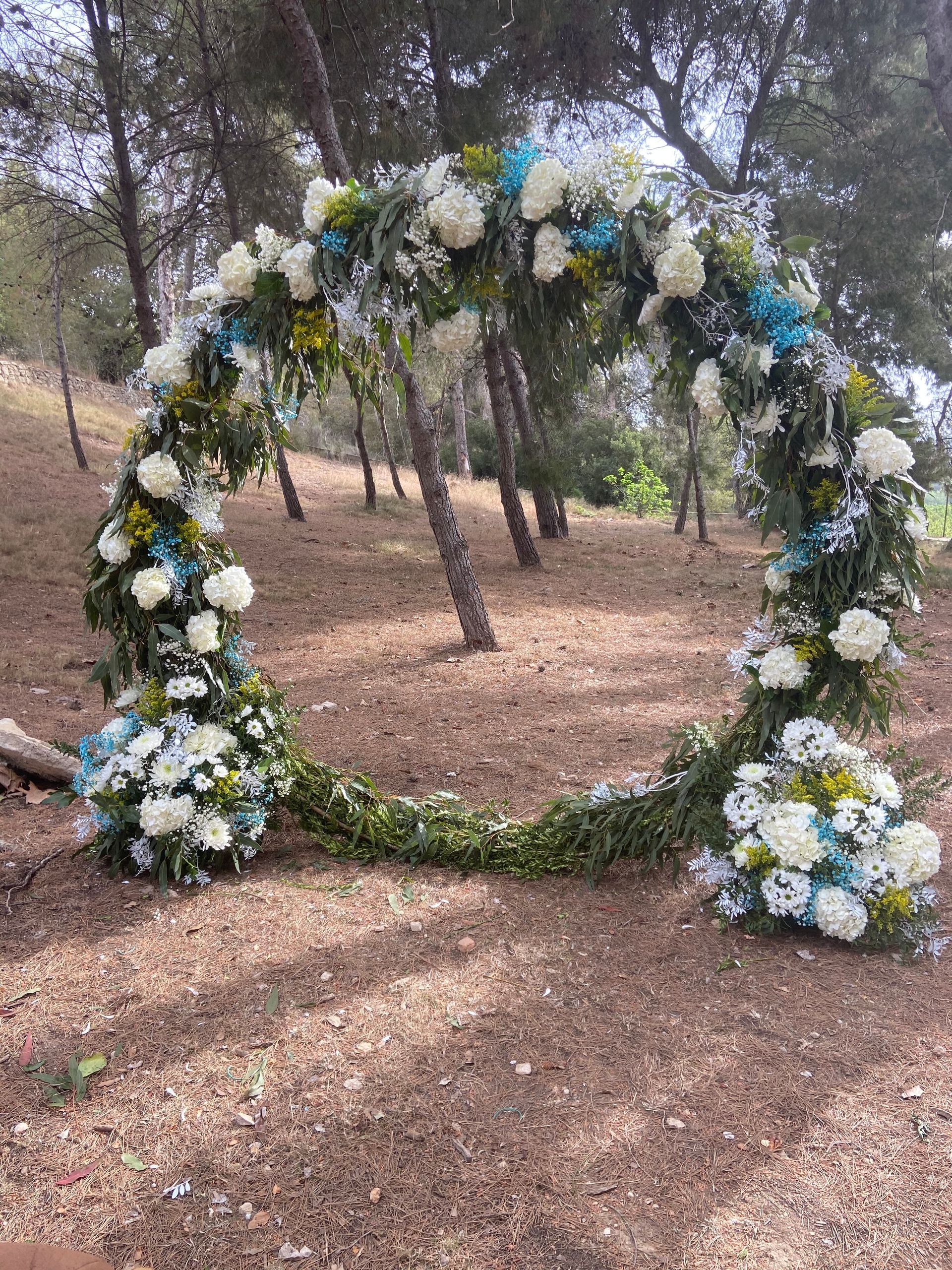Arco floral realizado con flores blancas y azules, situado en una zona boscosa.