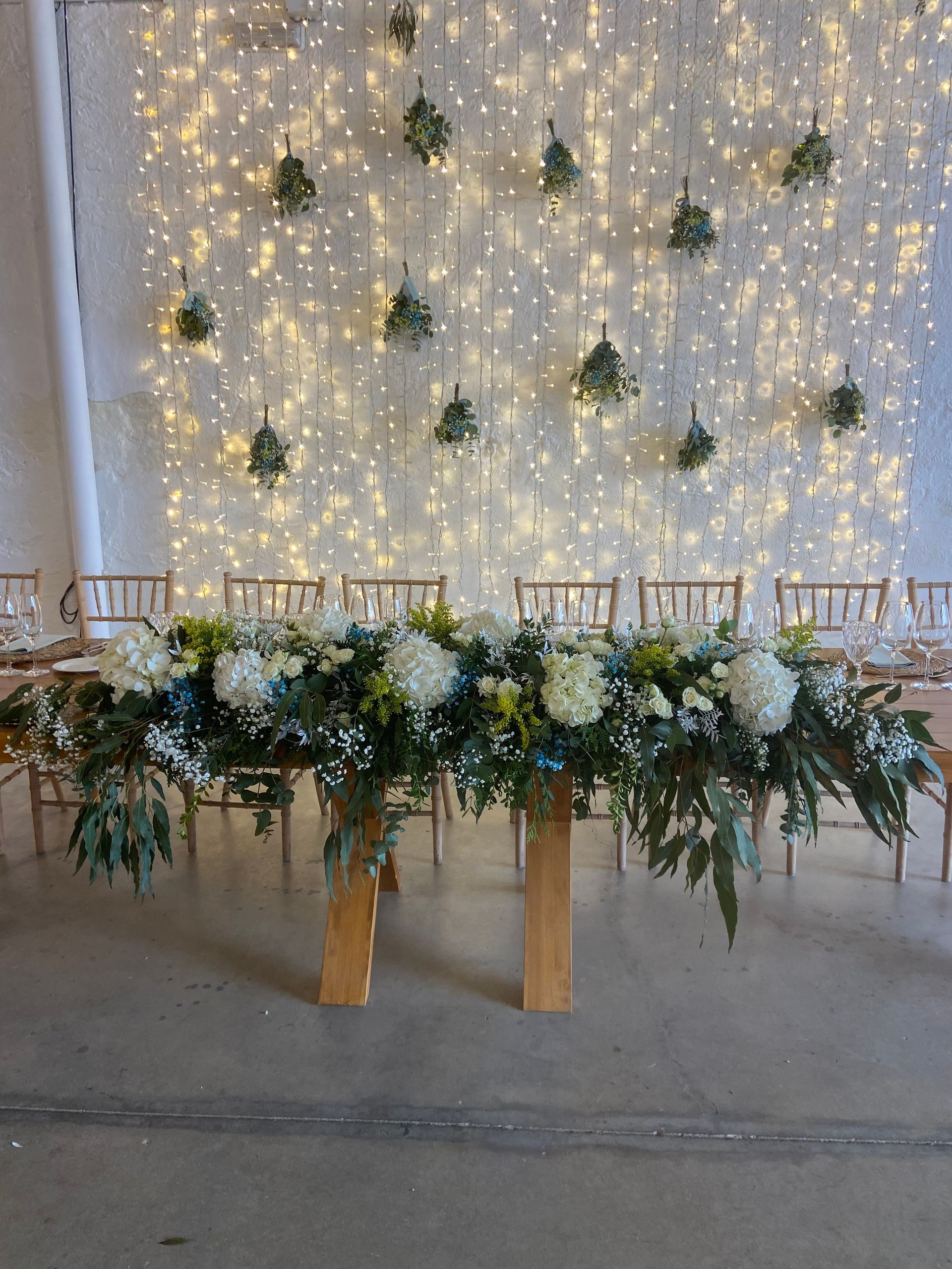Mesa rectangular larga con arreglo floral, frente a una pared con luces de cadena y vegetación colgante.