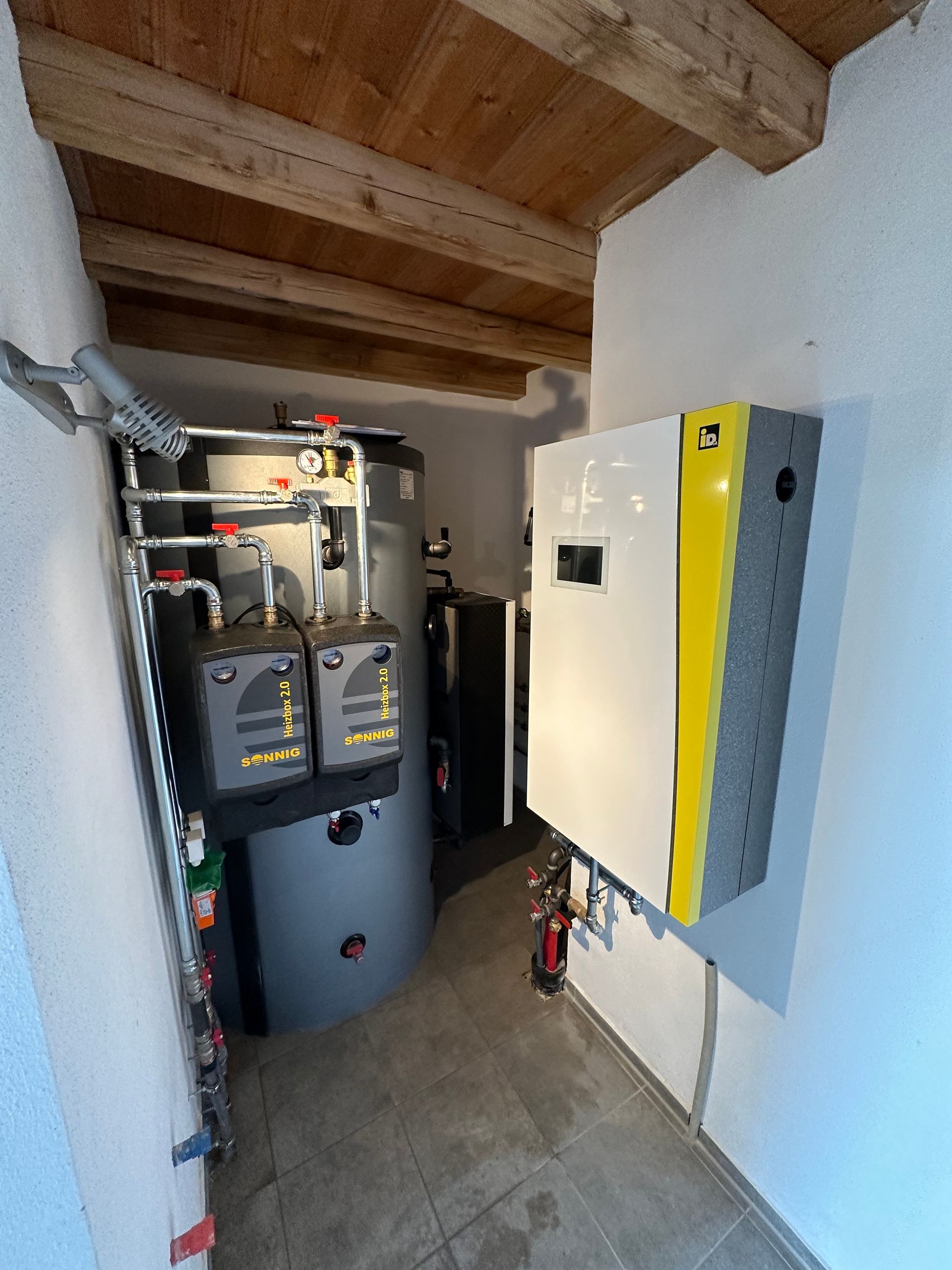 Ein kleiner Heizungsraum mit Wandgerät, Warmwasserspeicher und isolierten Rohrleitungen.