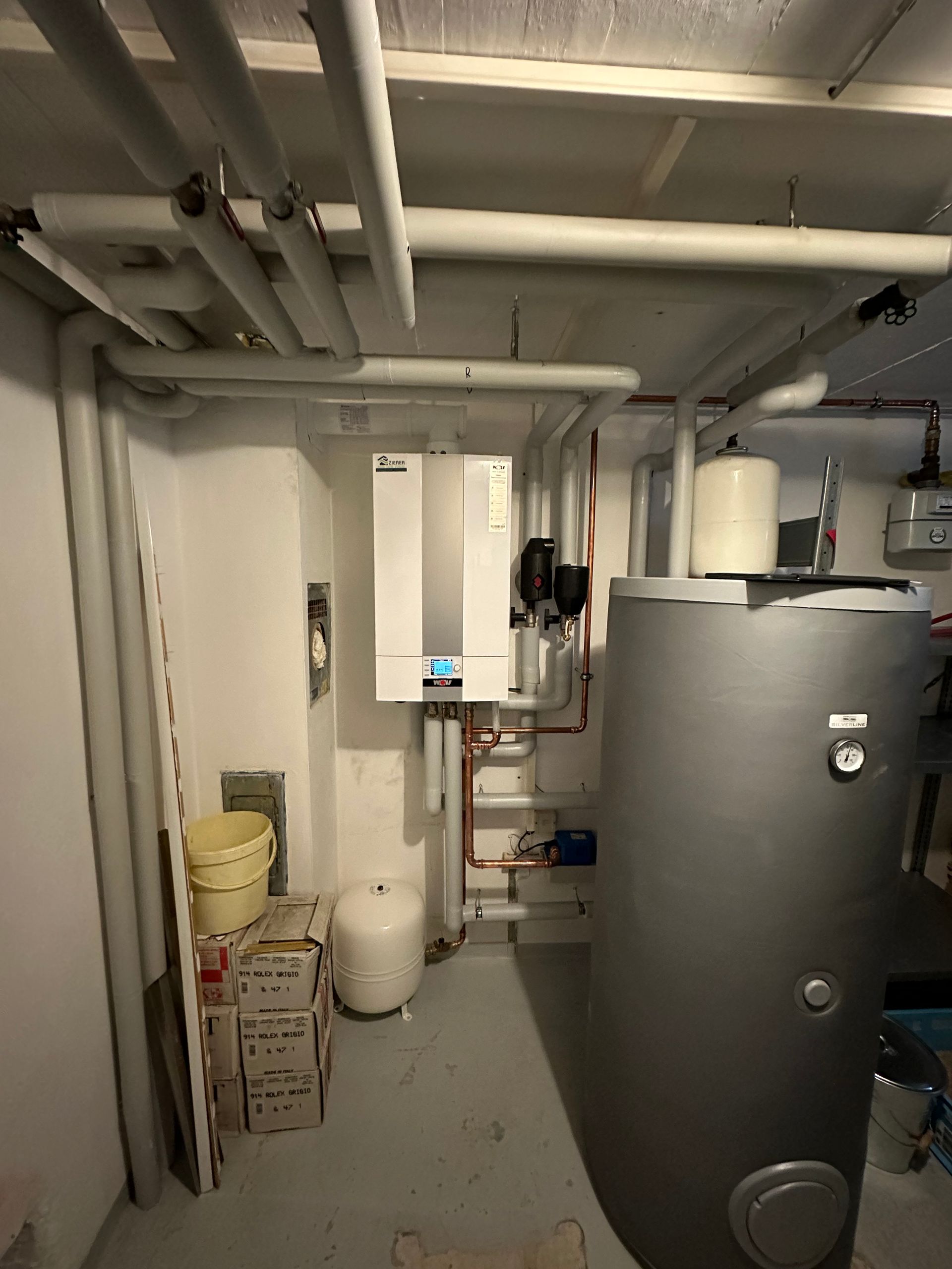 Heizungsraum mit Wandgerät, Warmwasserspeicher und isolierten Rohrleitungen.