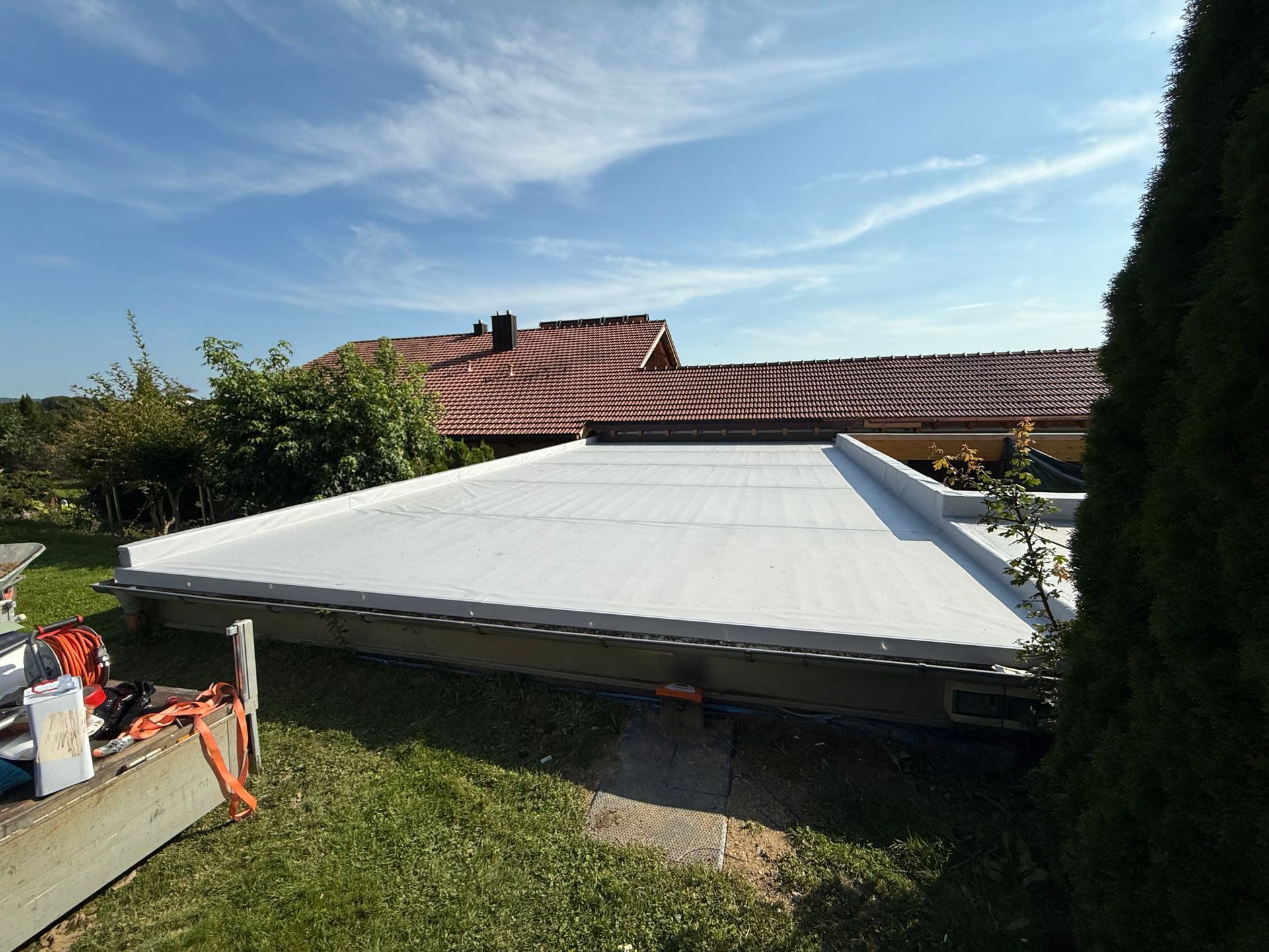Ein flaches Dach mit heller Abdichtung in einem Garten mit Bäumen und einem Wohnhaus im Hintergrund unter blauem Himmel.