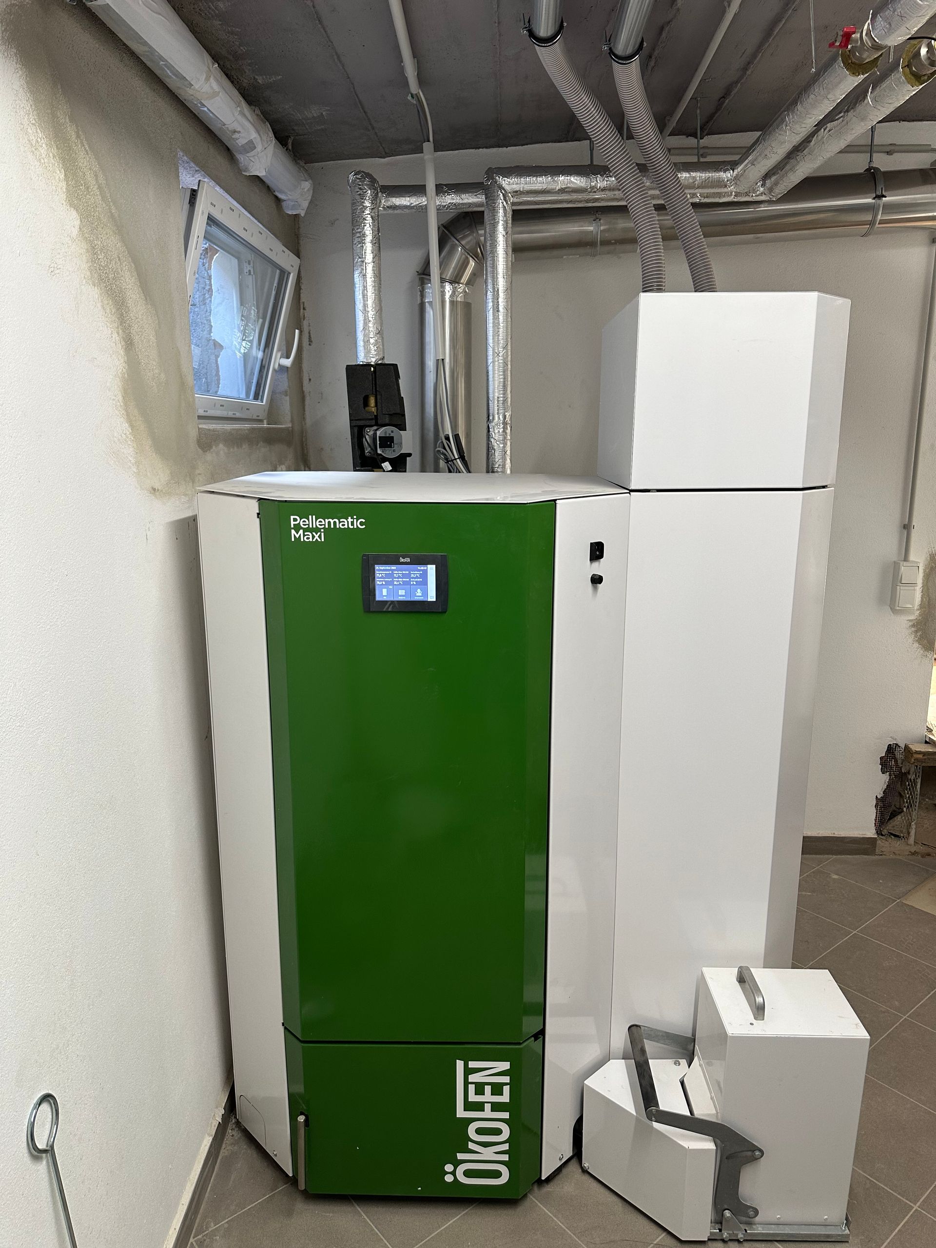 Ein moderner Pelletkessel in einem Heizraum mit grüner Front, Display und angeschlossenen Rohren sowie Fördereinheit.