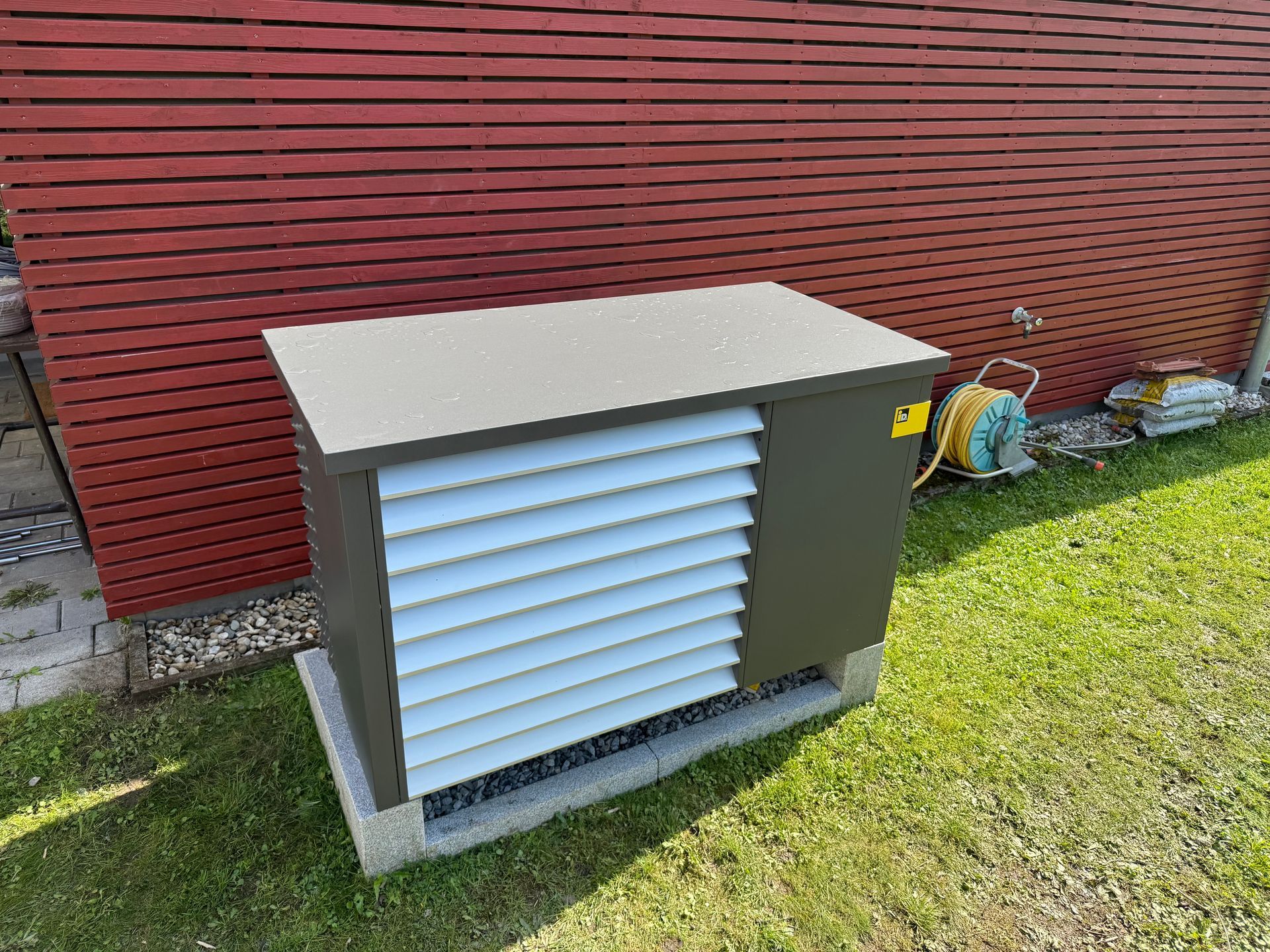 Moderne, wetterfeste Wärmepumpen-Außeneinheit im Garten neben Holzfassade.