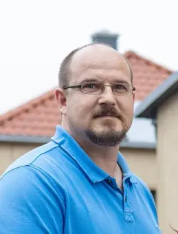 Ein Mann mit Brille und blauem Hemd steht vor einem Haus.