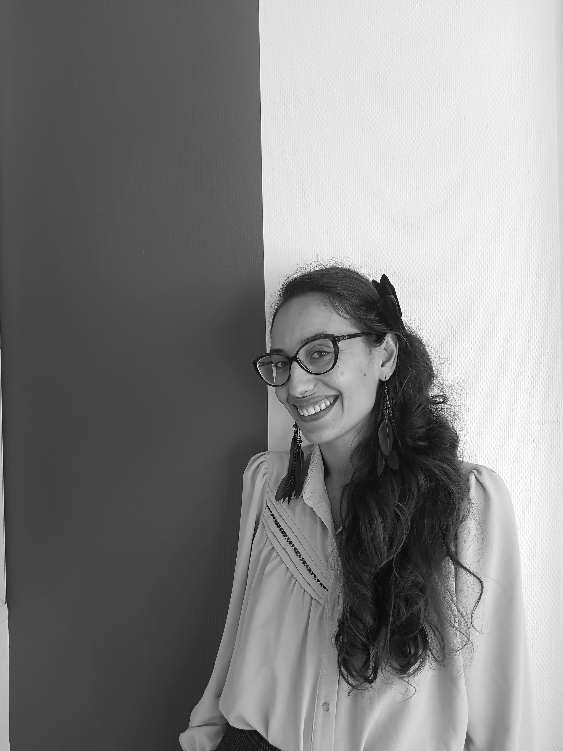 Une femme avec des lunettes sourit, appuyée contre un mur, de longs cheveux noirs.