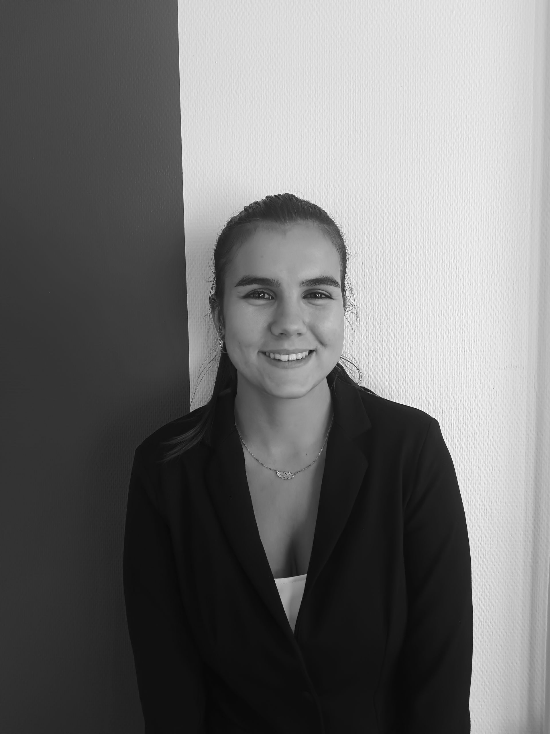 Une femme en blazer noir sourit, se tient devant un mur blanc et gris.