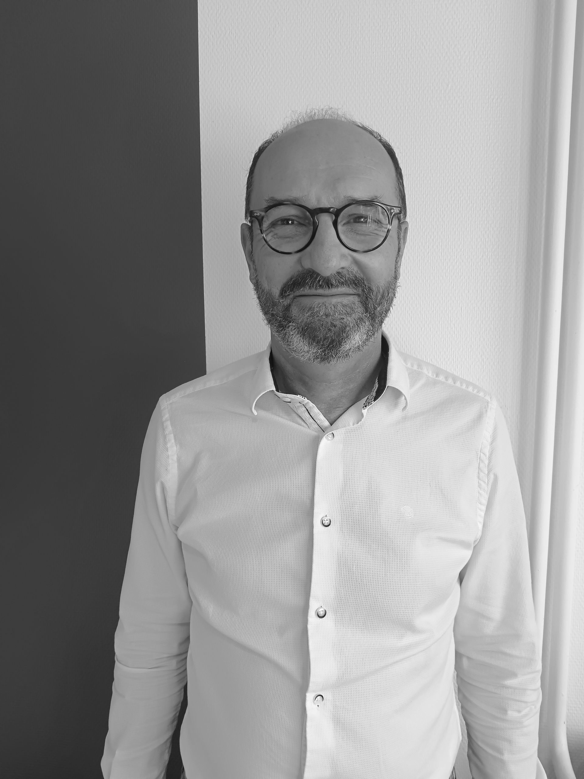 Homme avec des lunettes et une barbe, vêtu d'une chemise boutonnée, debout devant une fenêtre et un mur sombre.