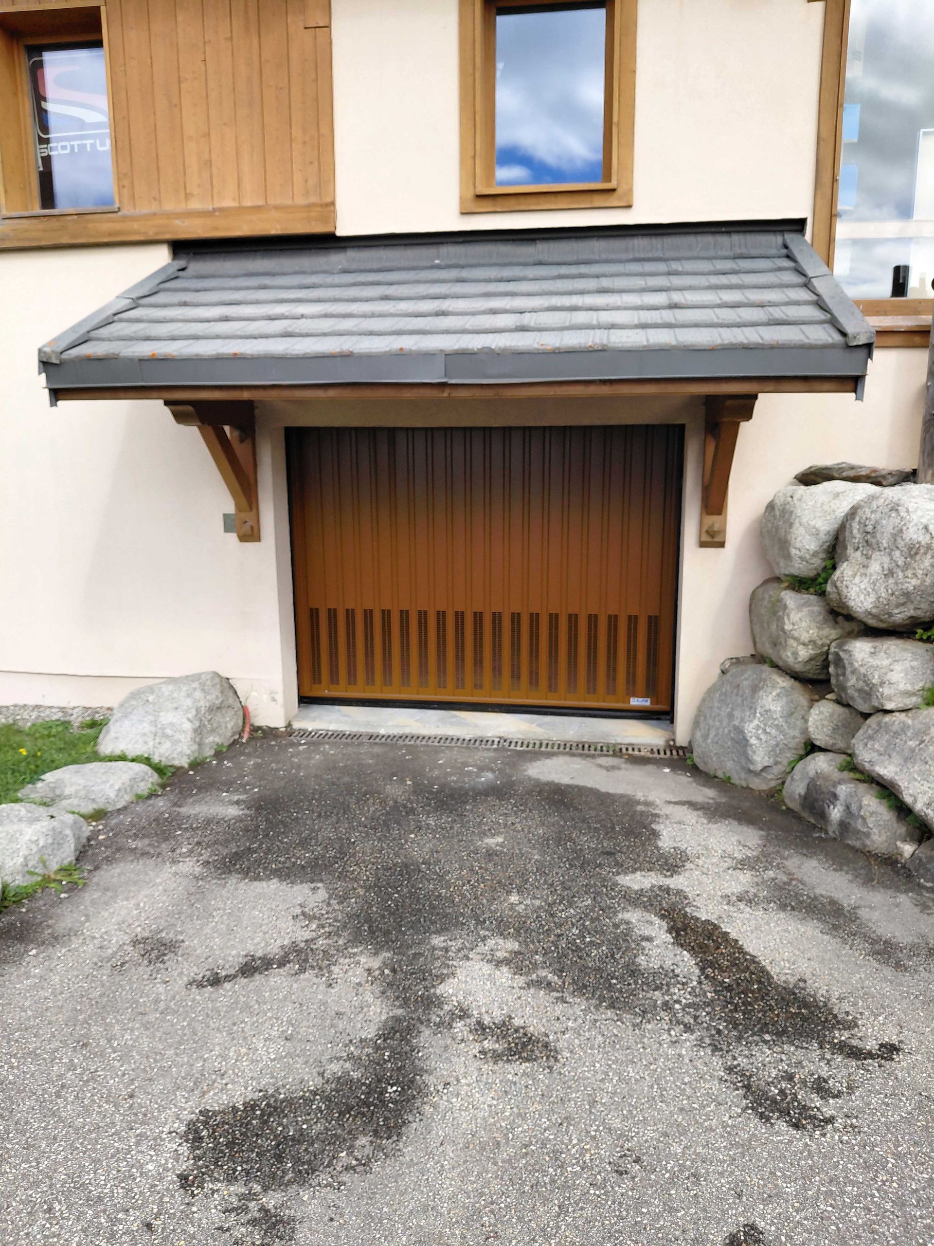Porte de garage marron sous un petit toit, allée en pierre.