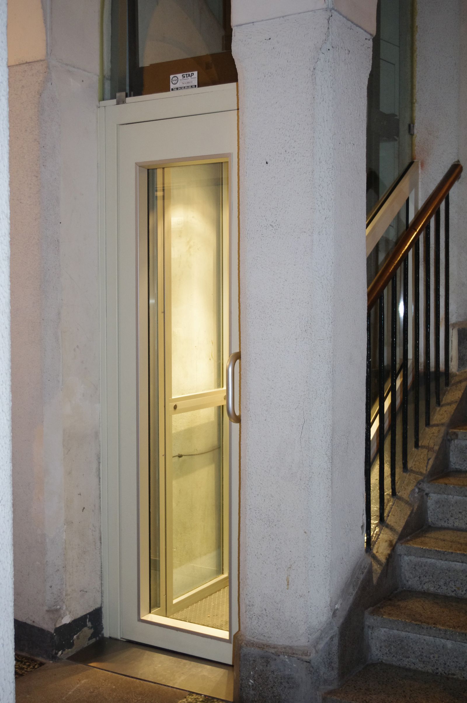 Ascenseur dans un immeuble, à côté d'un escalier. L'ascenseur a une porte vitrée. L'intérieur est éclairé.