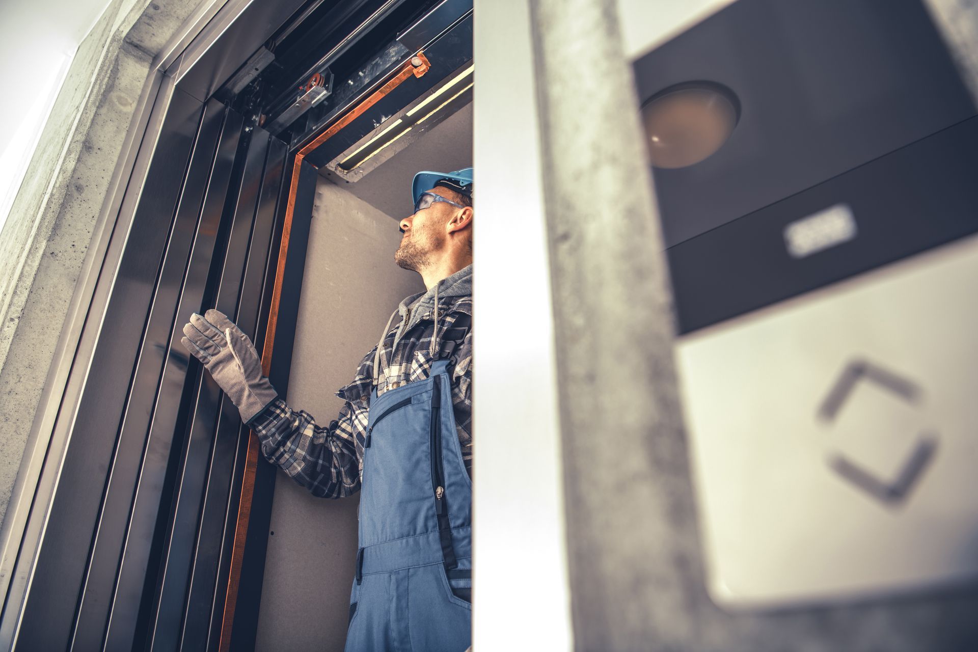 Un technicien d'ascenseur en salopette bleue inspecte les mécanismes de l'ascenseur à l'intérieur de la cage d'ascenseur.