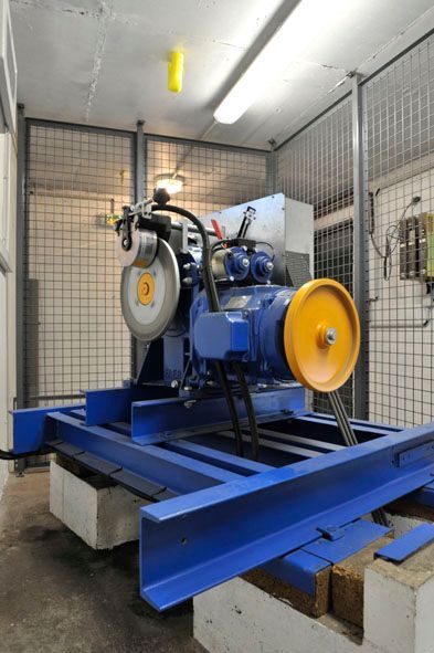 Moteur d'ascenseur bleu avec poulies jaunes, monté sur un châssis métallique bleu dans une pièce fermée.