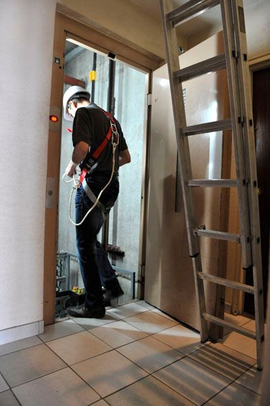 Un technicien d'ascenseur se trouve dans l'embrasure de la porte, portant un harnais et un casque, avec une échelle à côté.