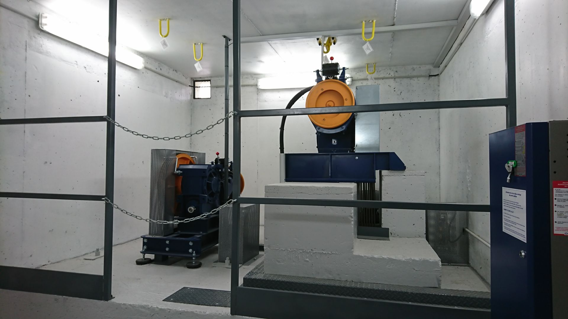 Mécanisme d'ascenseur à l'intérieur d'une pièce, derrière une grille métallique. Il comprend une grande roue, un moteur et un panneau de commande.