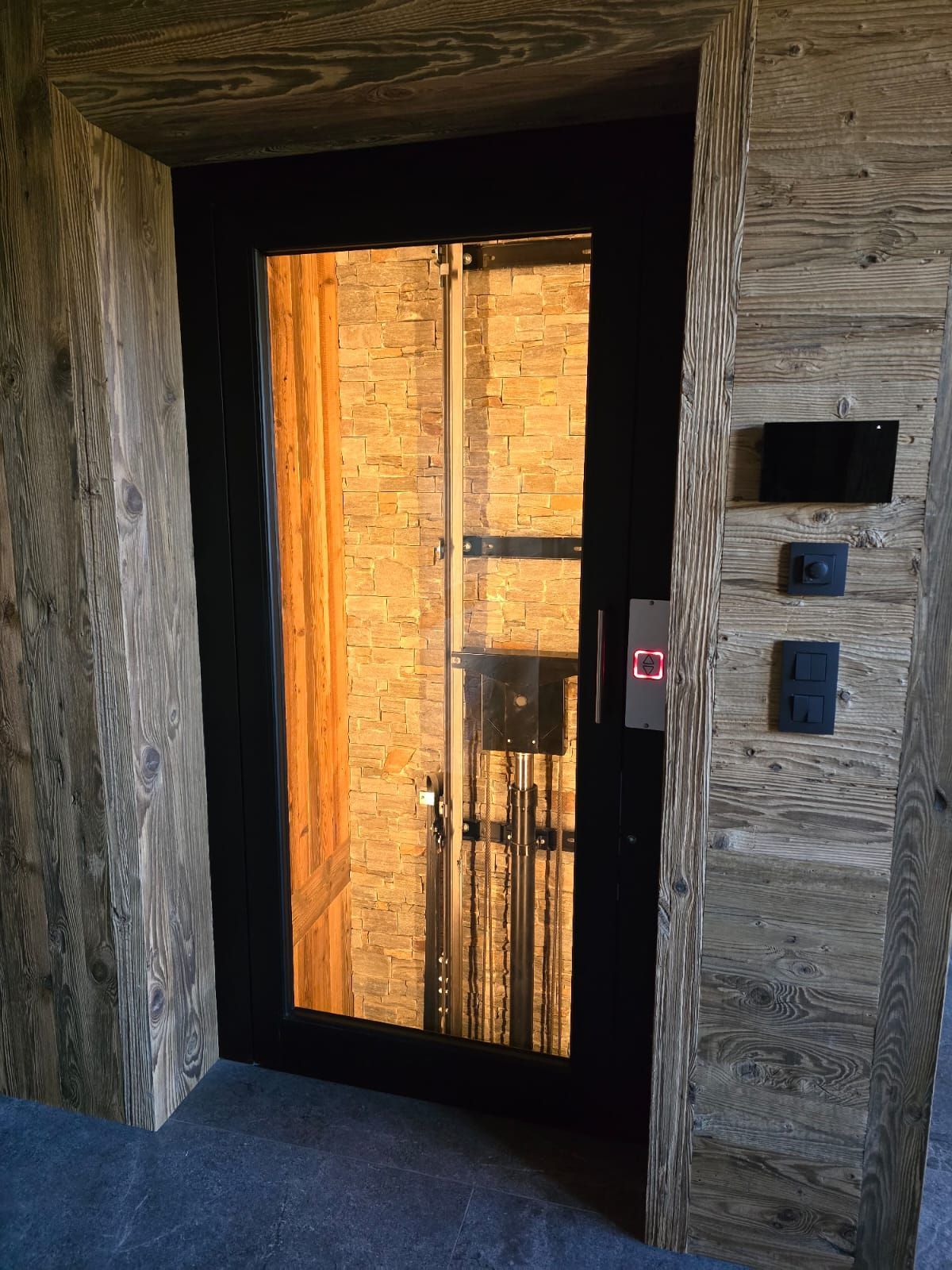 Ascenseur à porte vitrée, intérieur à cadre en bois. Bouton rouge illuminé. Panneau de commande à droite.
