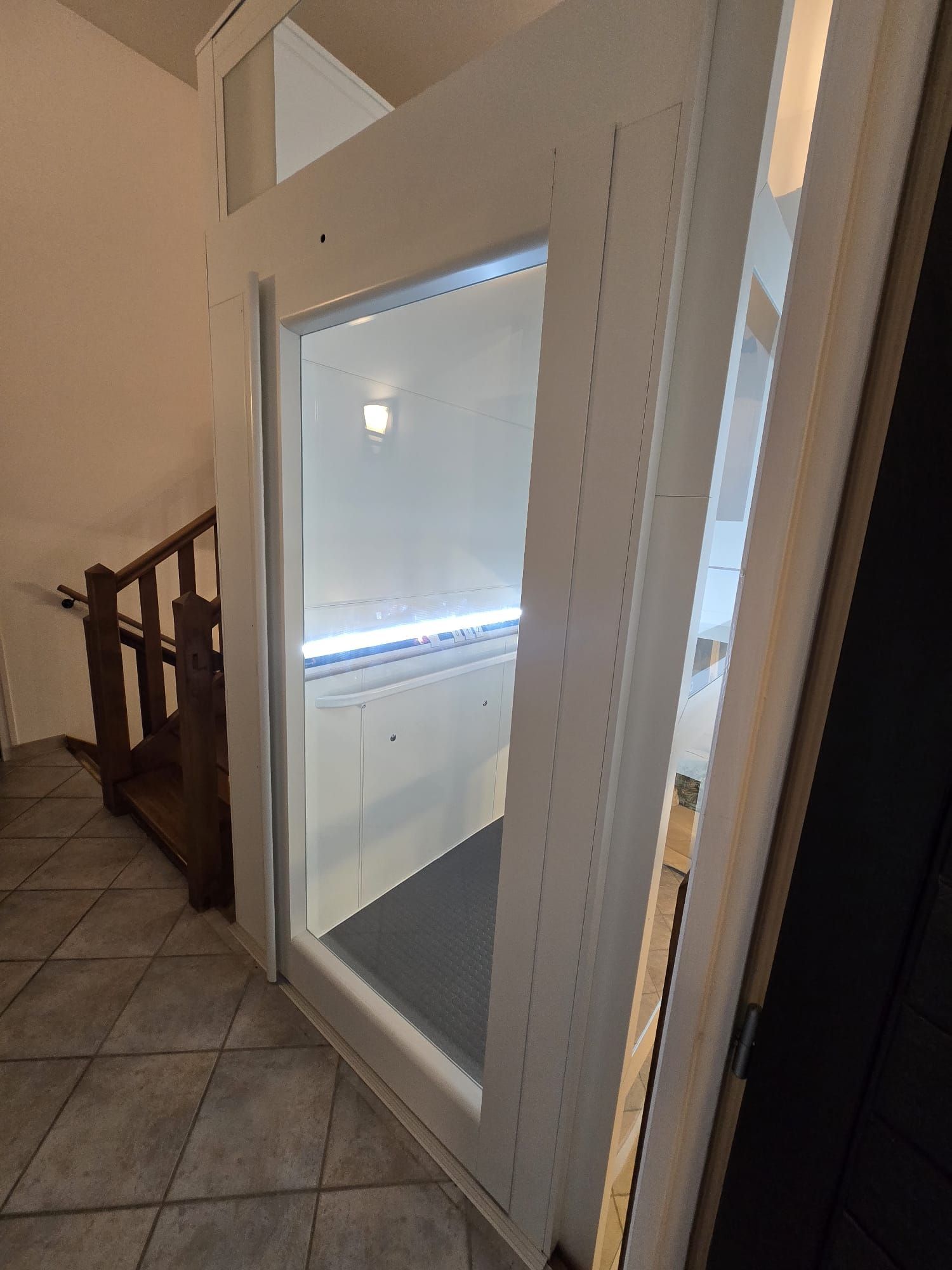 Ascenseur blanc avec porte vitrée dans une maison, à côté d'un escalier en bois.