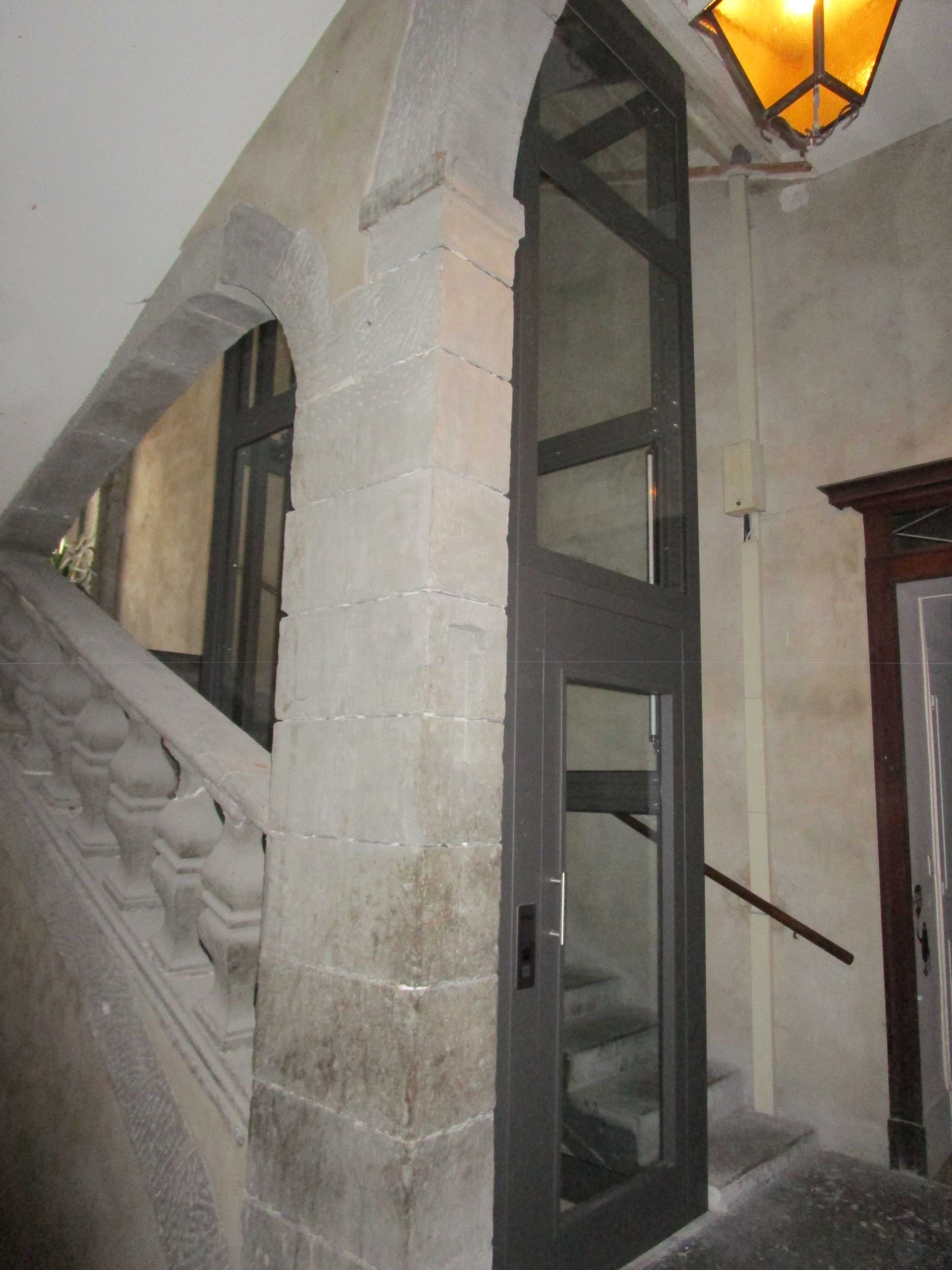 Un ascenseur aux portes vitrées jouxte un escalier dans un immeuble en pierre.
