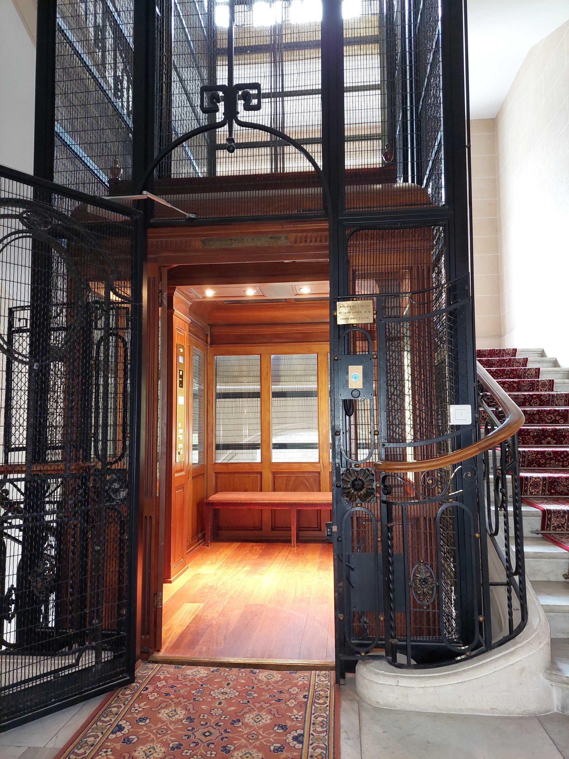 Ascenseur orné avec portes en fer forgé ajourées, intérieur en bois et petite banquette. Un tapis rouge mène à l'escalier.