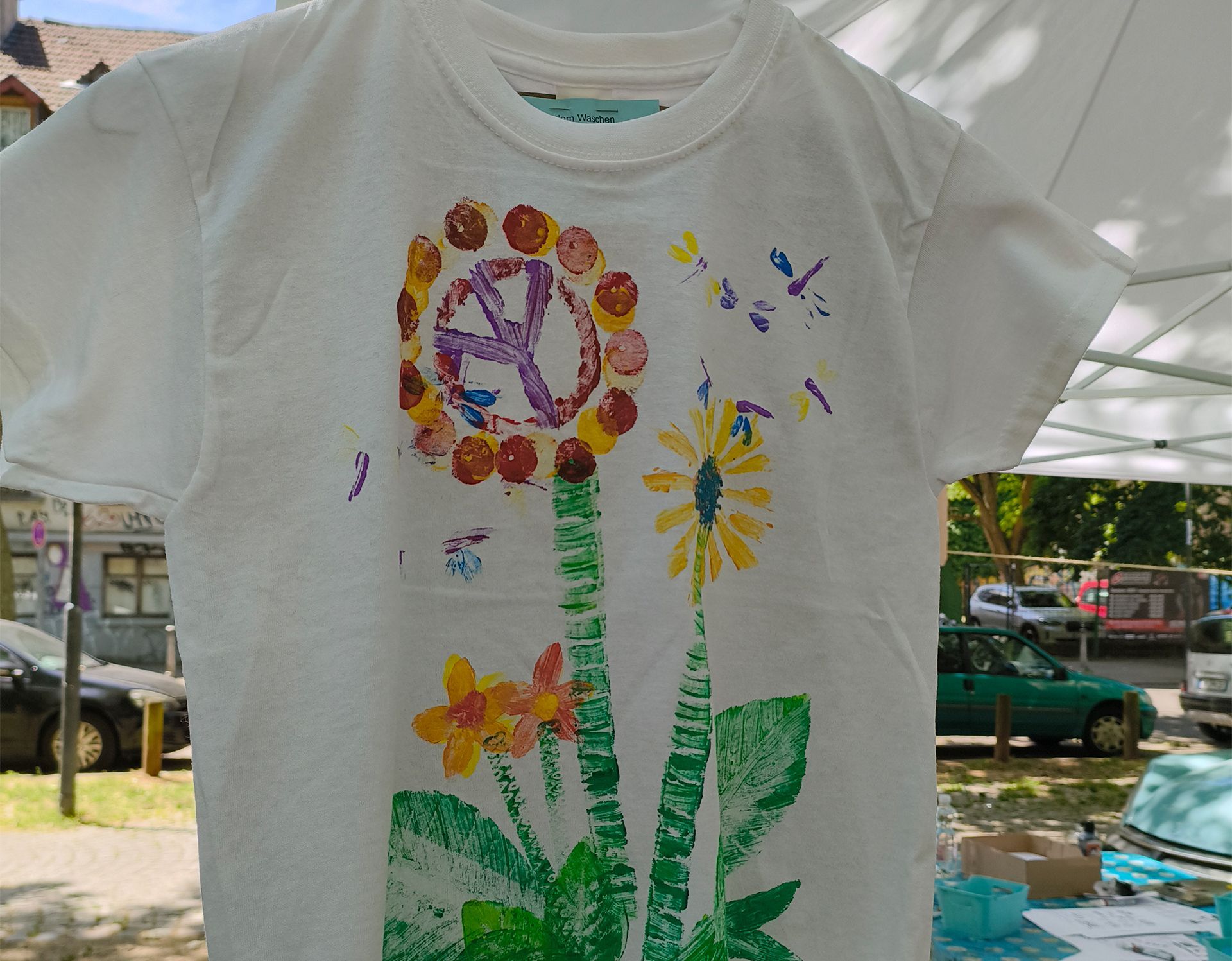 Weißes T-Shirt mit bunten, handgemalten Blumen, die draußen in einem sonnigen Garten hängen.