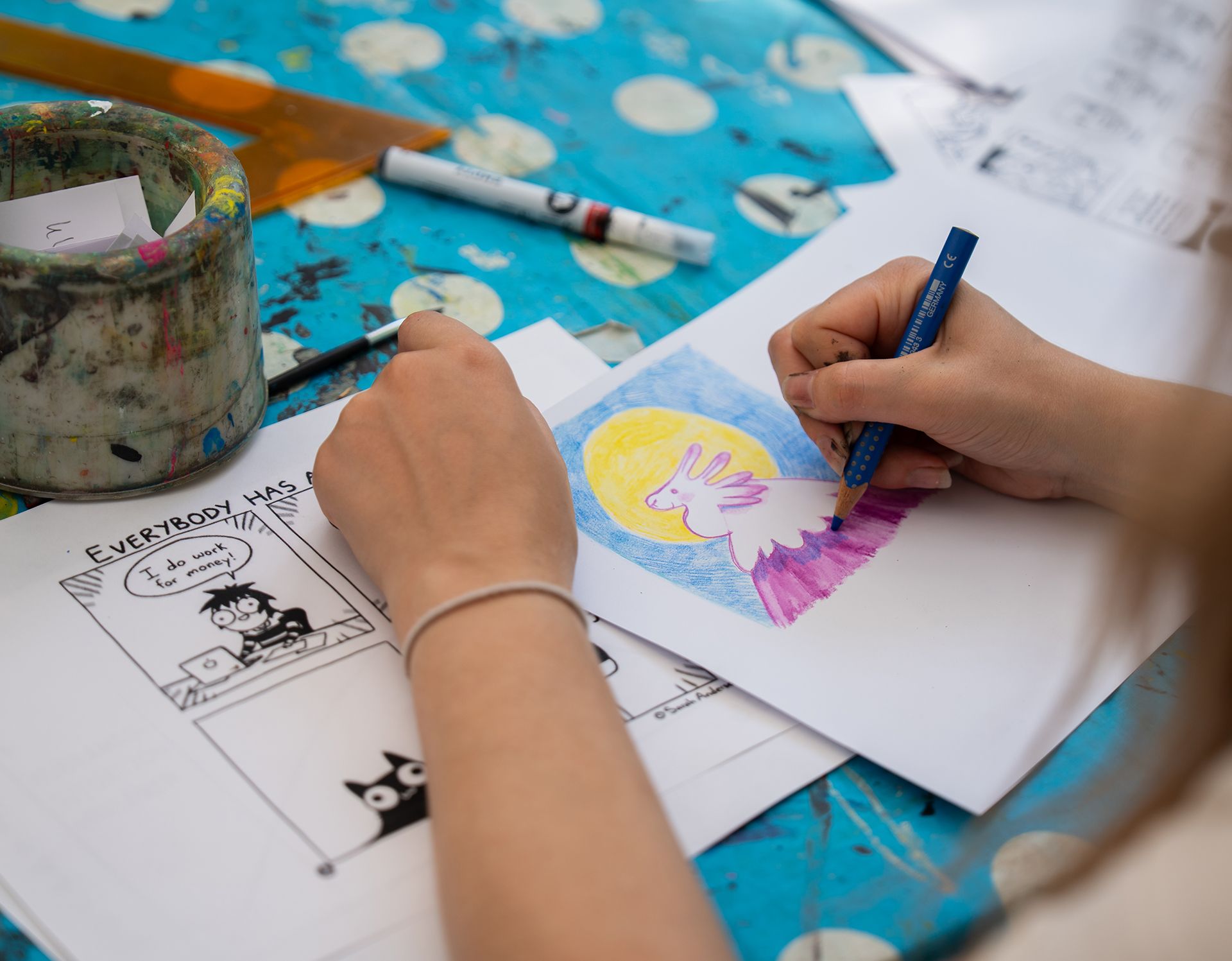 Hände malen eine Zeichnung auf Papier an einem Basteltisch mit Farbe und Stiften in der Nähe.