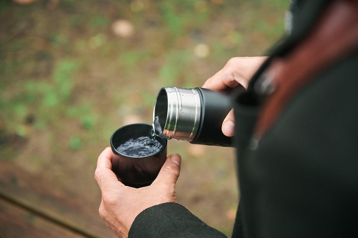 Hands pouring water from a metal thermos into a black cup outdoors. Dopper Water Bottle.
