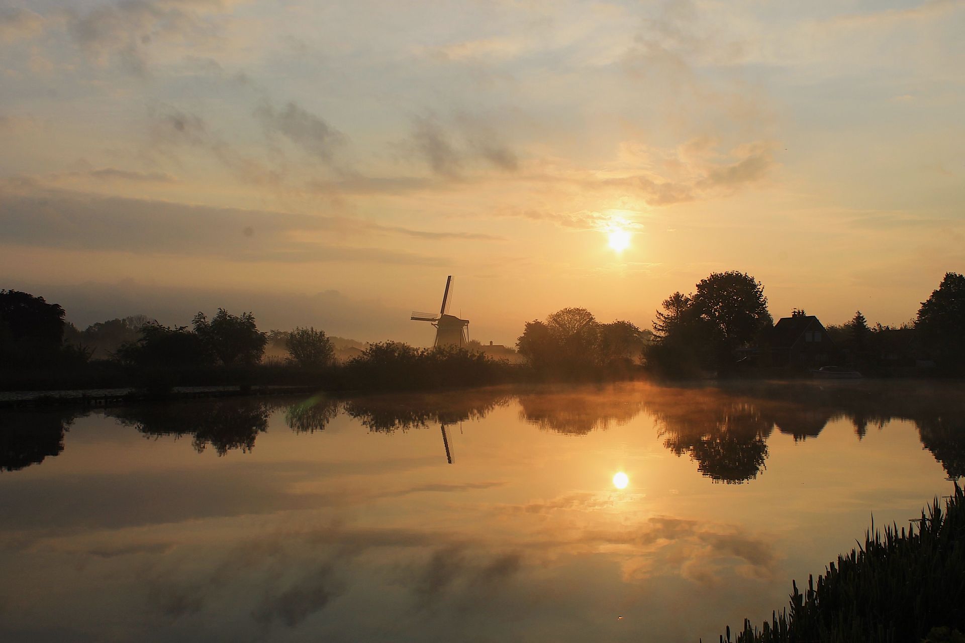 Sunset over a calm river with silhouetted trees and a windmill reflected in the water.