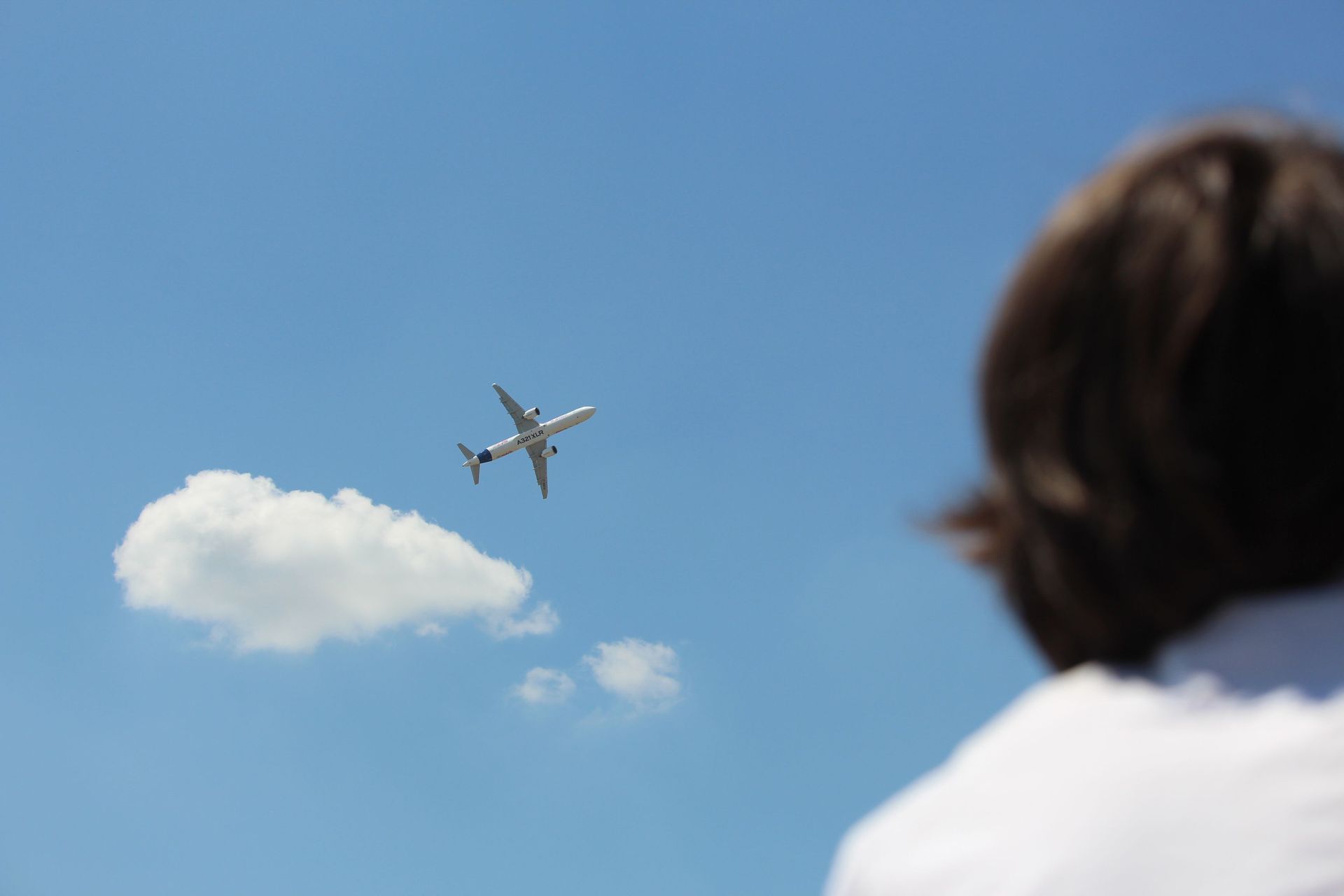 Airplane flying across a blue sky above a white cloud, viewed past a person’s shoulder.